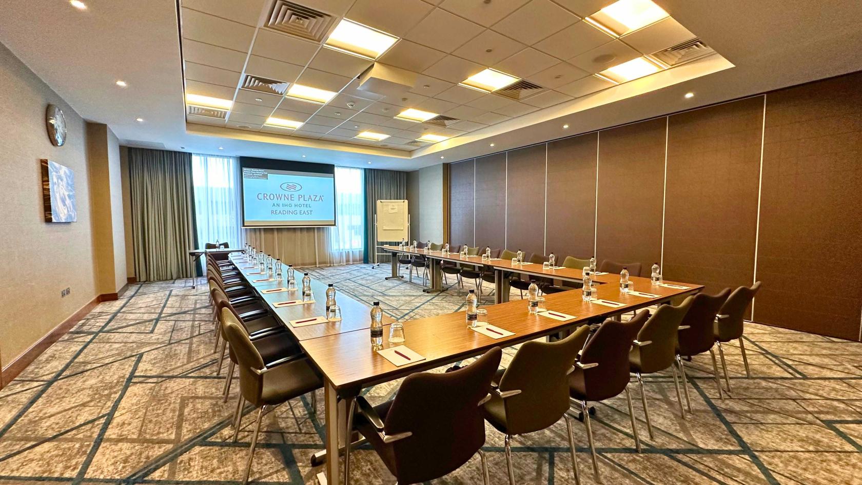 An auditorium with U-shaped tables, bottled water, notepads, and a projector screen at Crowne Plaza Reading Windsor Room.
