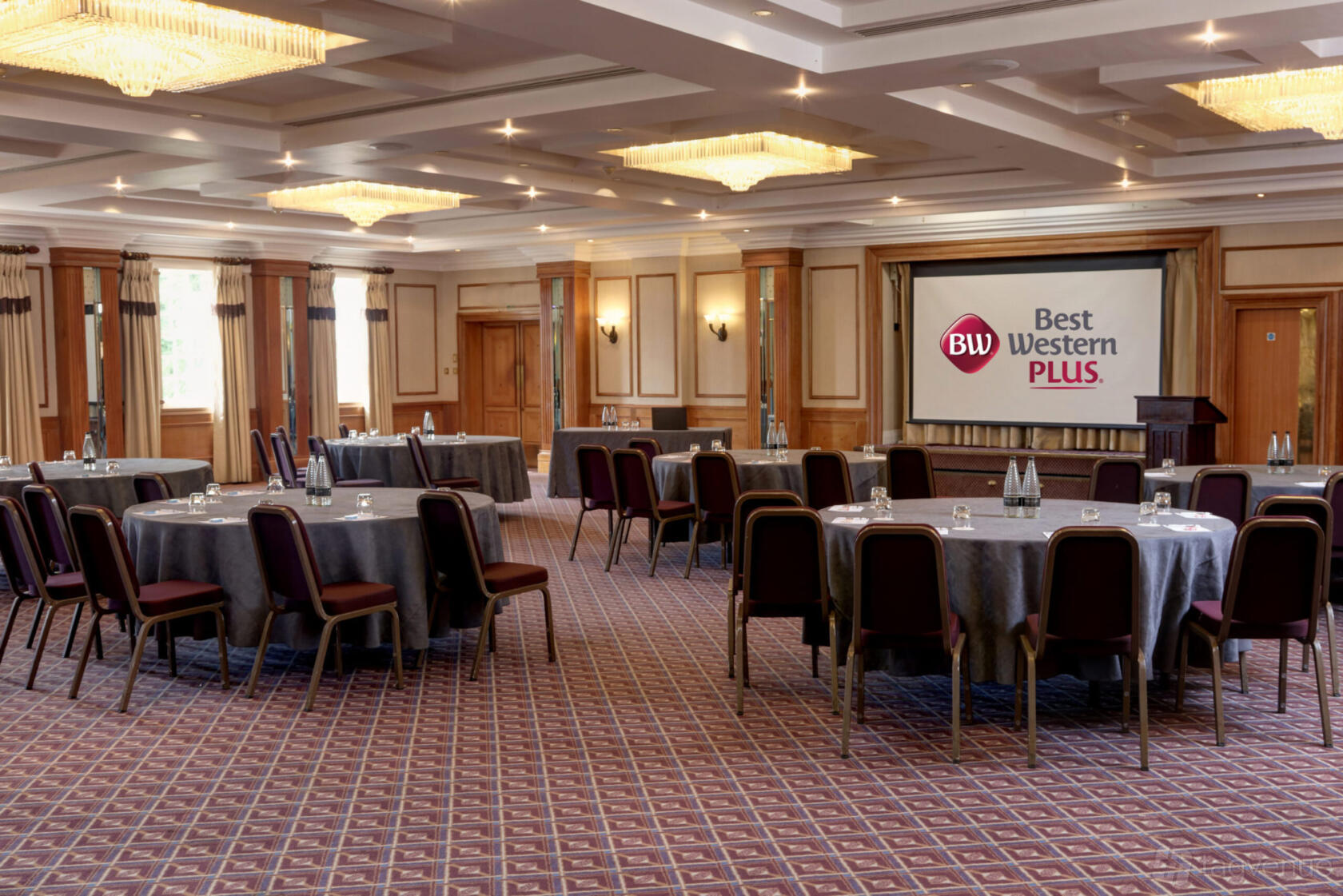 An event space with round tables, cushioned chairs, chandeliers, and a large projector screen at Best Western Plus Manor Hotel.