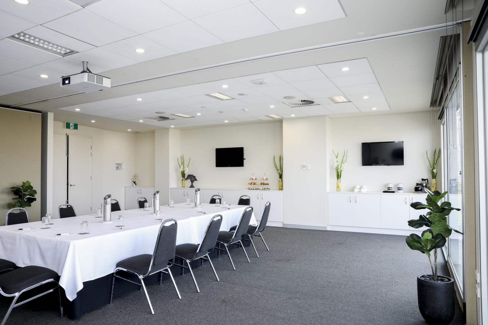 A meeting room with a U-shaped table setup, white linens, wall-mounted screens, and potted plants at Melton Entertainment Park.
