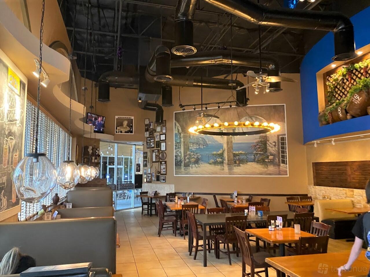 A restaurant with wooden tables, booth seating, pendant lighting, and a large Greece mural at Opa! Greek Taverna.