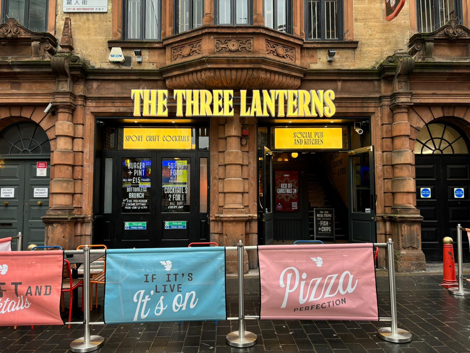 A pub with ornate stone facade, bright yellow signage, and outdoor banners at The Three Lanterns.