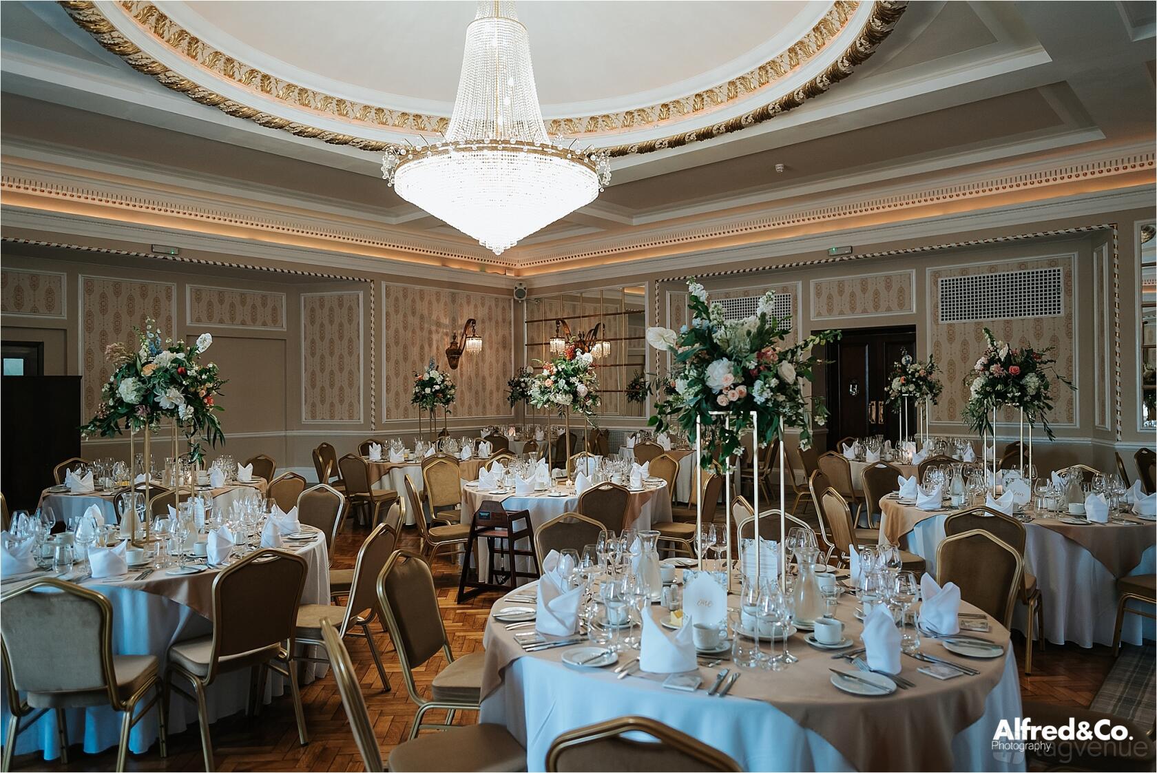 A function room with round tables set with white linens, floral centerpieces, and a large chandelier at Manchester Hall