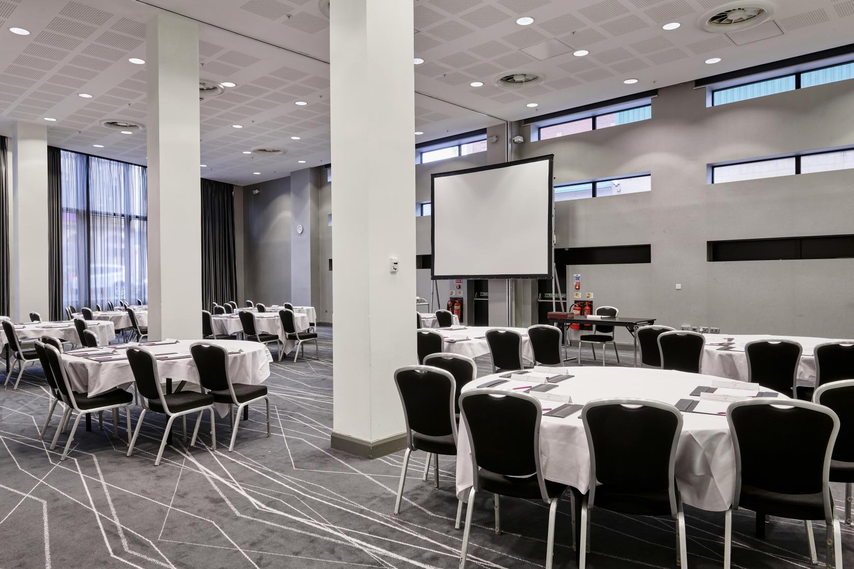 A hotel hall with round tables, black chairs, floor-to-ceiling windows, and a projection screen at Crowne Plaza Manchester City Centre.
