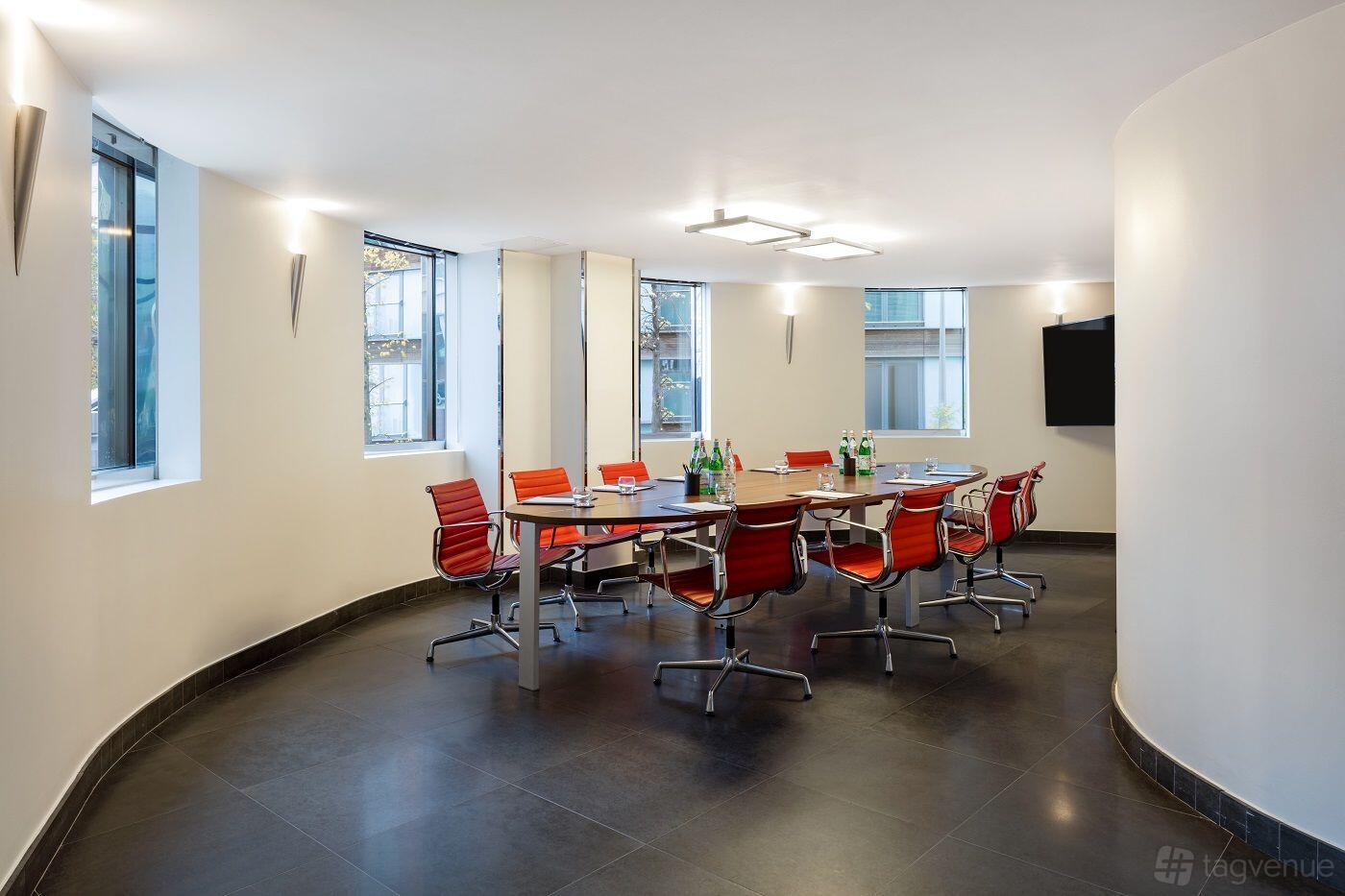A meeting room with red chairs, a large rectangular table, wall-mounted screen, and floor-to-ceiling windows at Radisson Blu Hotel London Canary Wharf East.
