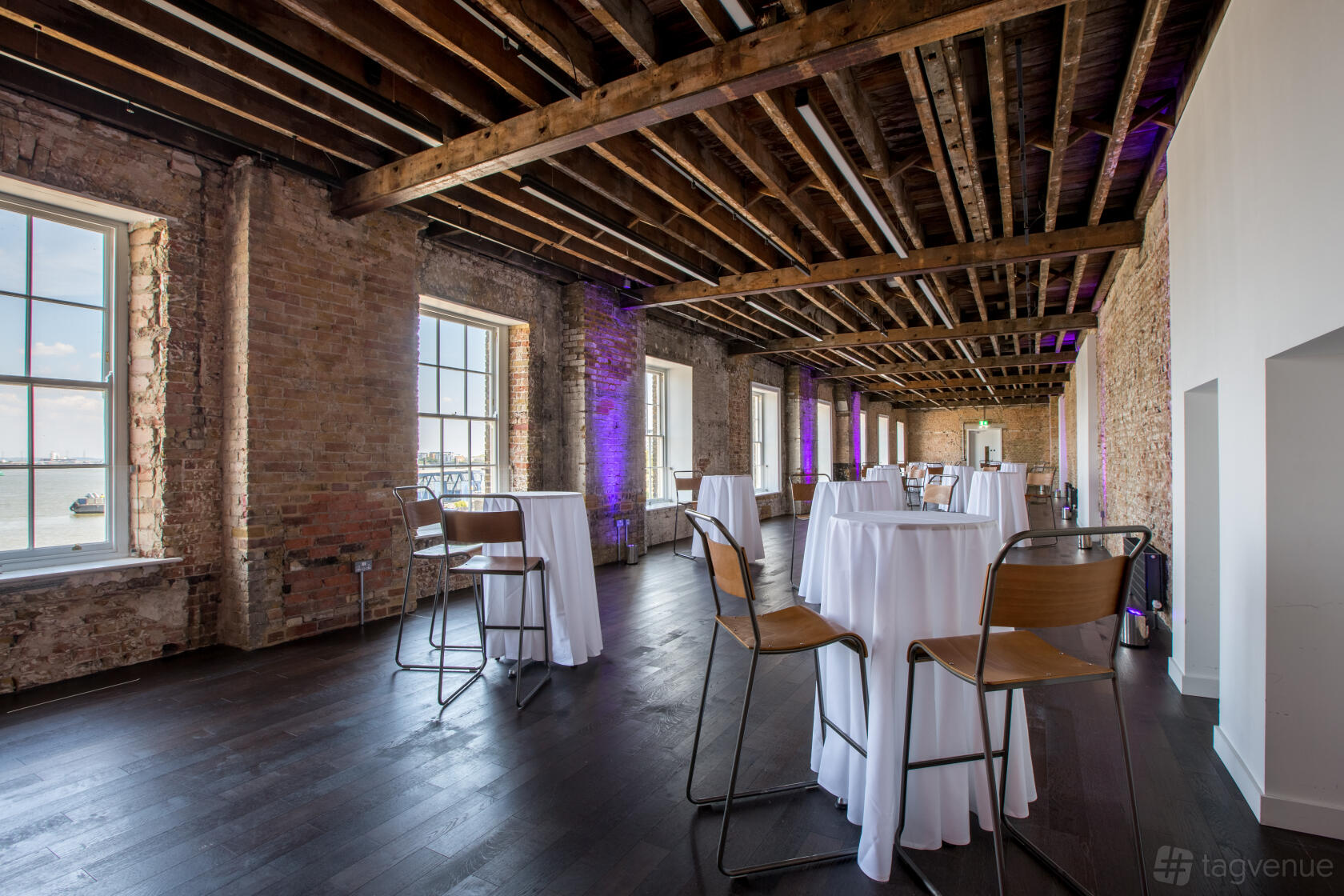 An event venue with exposed brick walls, wooden ceiling beams, tall windows and cocktail tables at Woolwich Works.