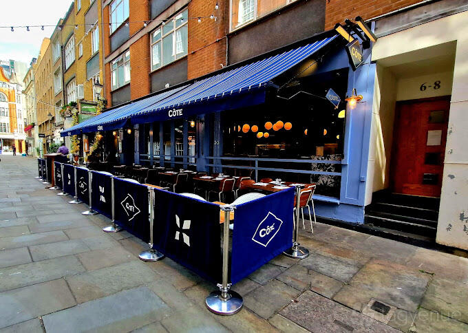An outdoor restaurant patio with tables, chairs, blue awning, and branded barriers at Cote Brasserie St. Christopher's Place.