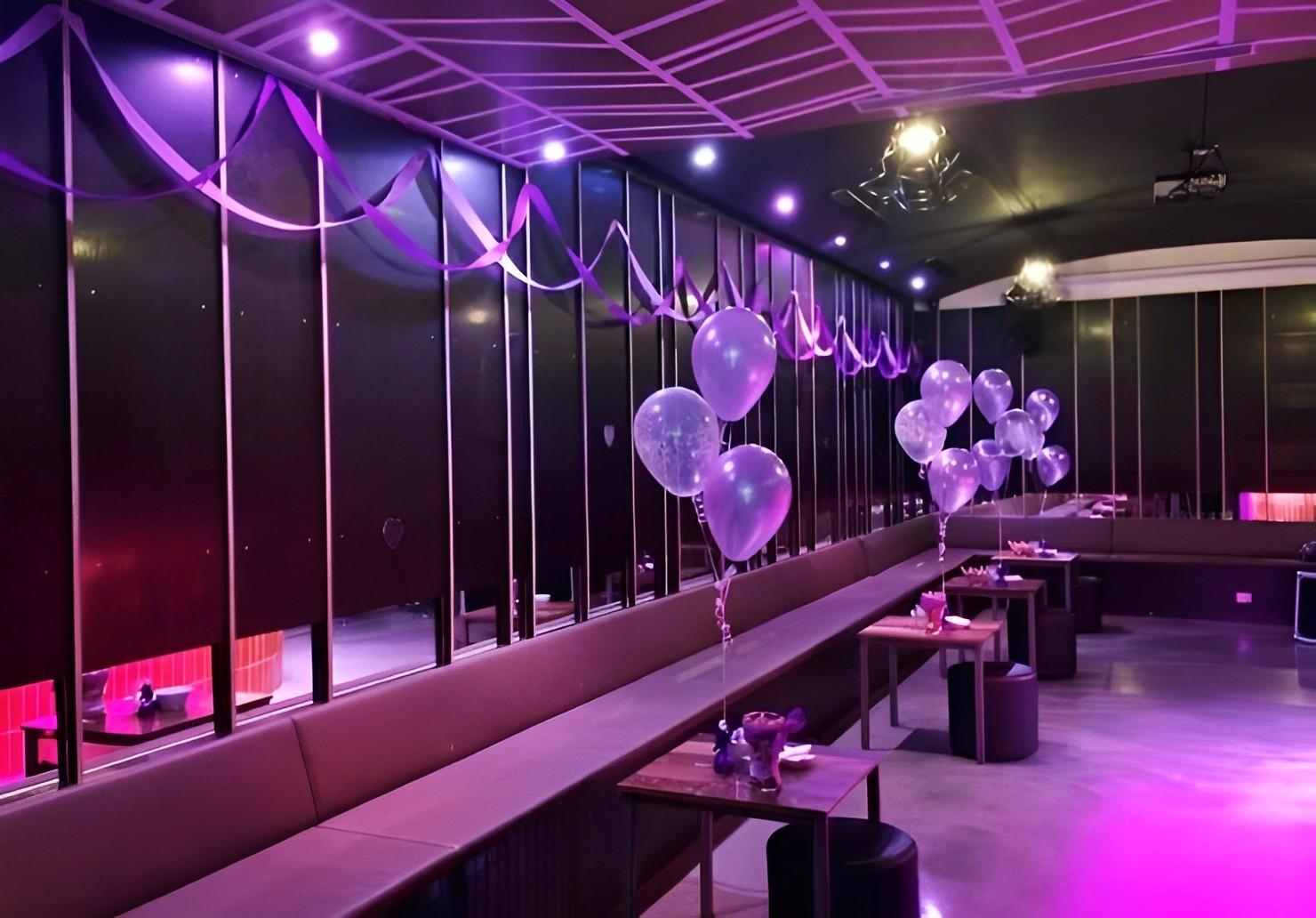 A lounge with purple lighting, long banquette seating, and tables decorated with balloon bouquets at Arcadia Hotel.