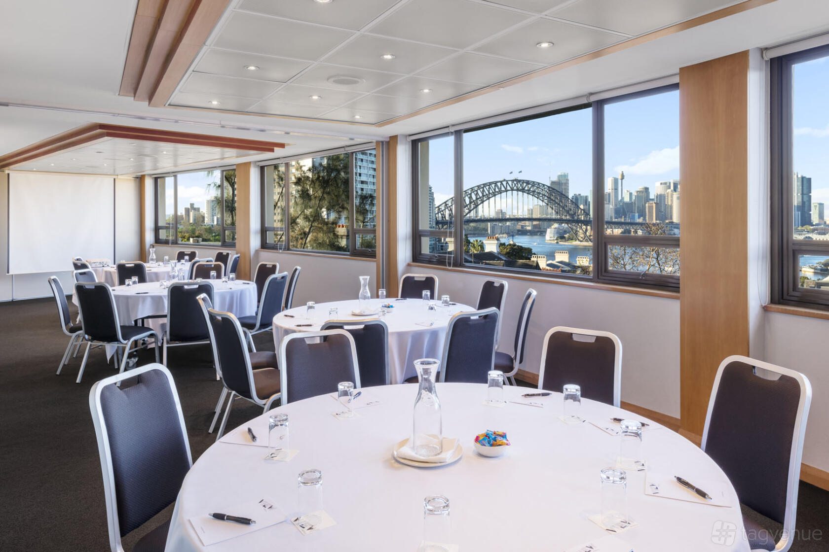 A meeting room with round tables, black chairs, large windows with city and harbor views at View Sydney.