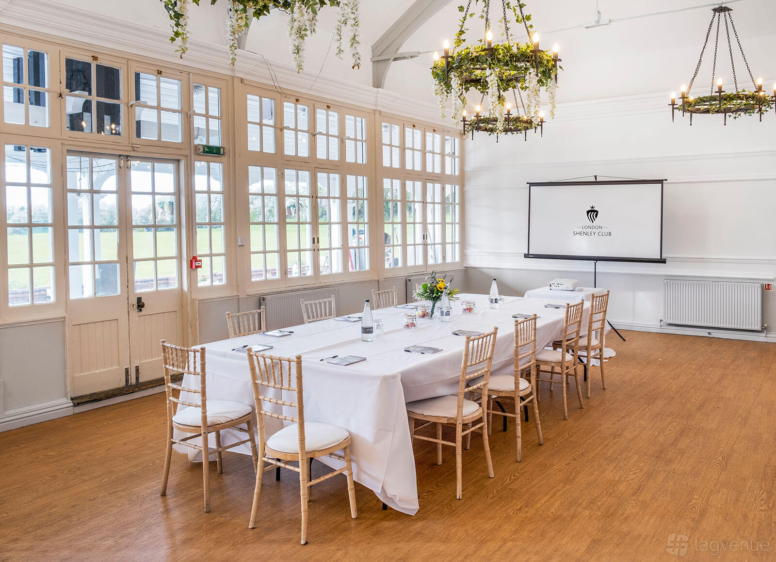 A bright hall with large windows, hanging greenery, and a U-shaped table with white linens at London Shenley Club.