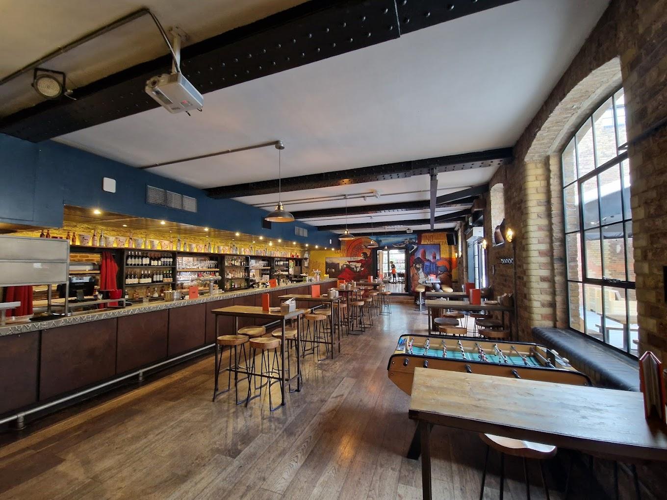 A bar in a restaurant with exposed brick walls, high tables, a foosball table, and large arched windows at Camino King's Cross.