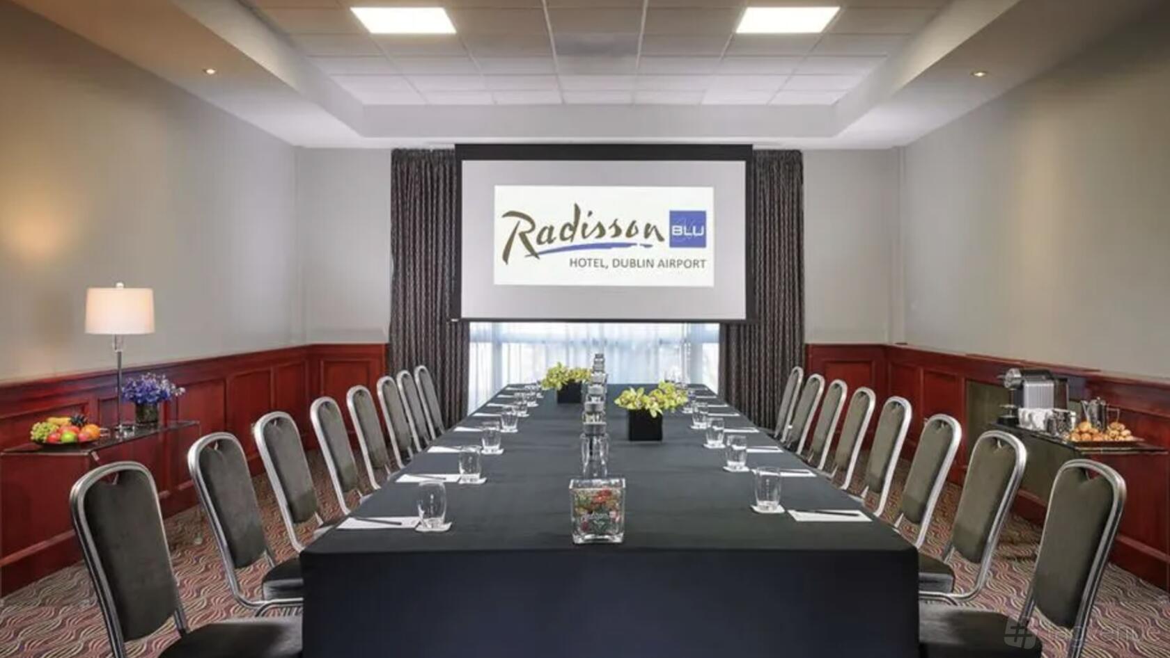 A hotel meeting room with U-shaped tables, black chairs, a projector screen, and floral centerpieces at Radisson Blu Hotel, Dublin Airport.