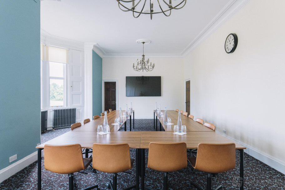 A meeting room with a U-shaped wood table, tan chairs, chandeliers, and a wall clock at Ashridge House.