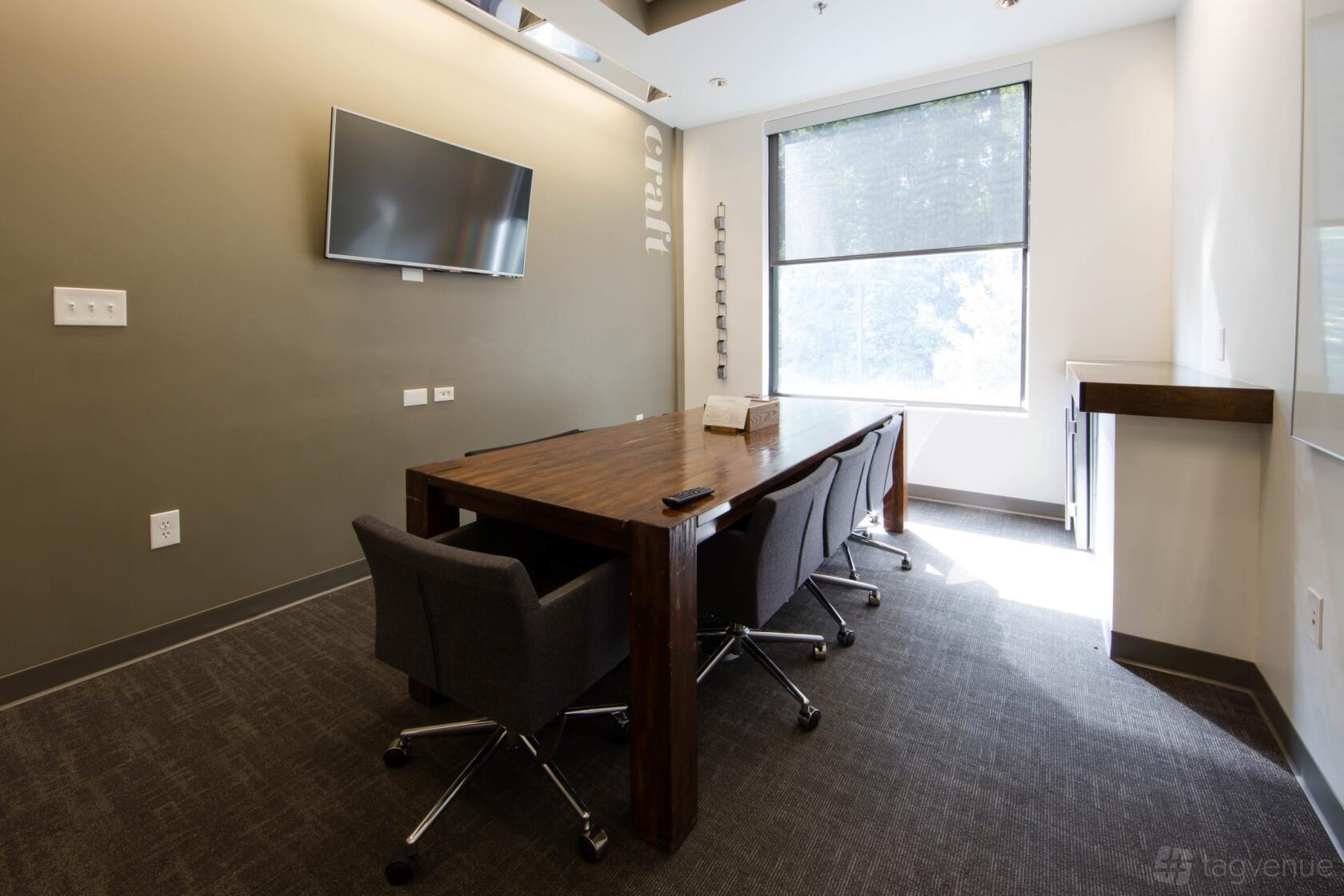 A meeting room with a wooden conference table, four gray chairs, wall-mounted TV, and large window at ROAM ALPHARETTA.