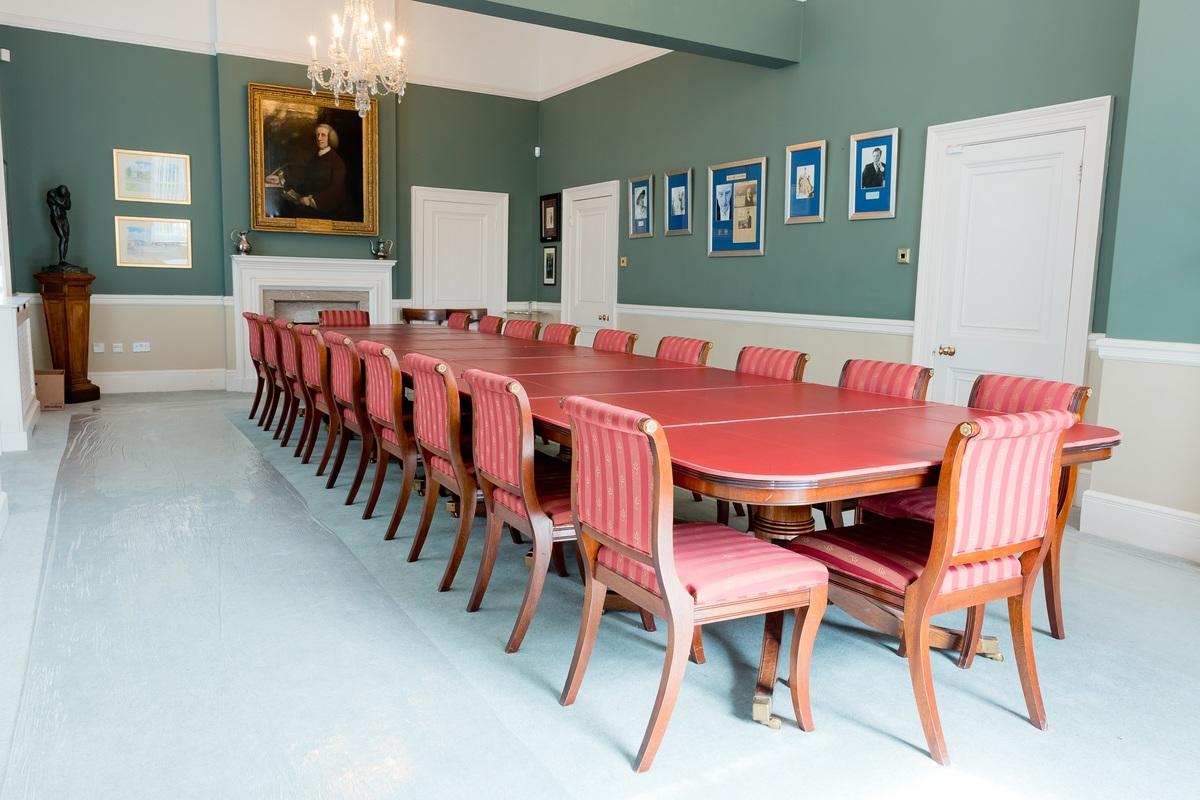 A conference room with a long red meeting table, striped chairs, framed portraits, and a chandelier at Mill Hill School.