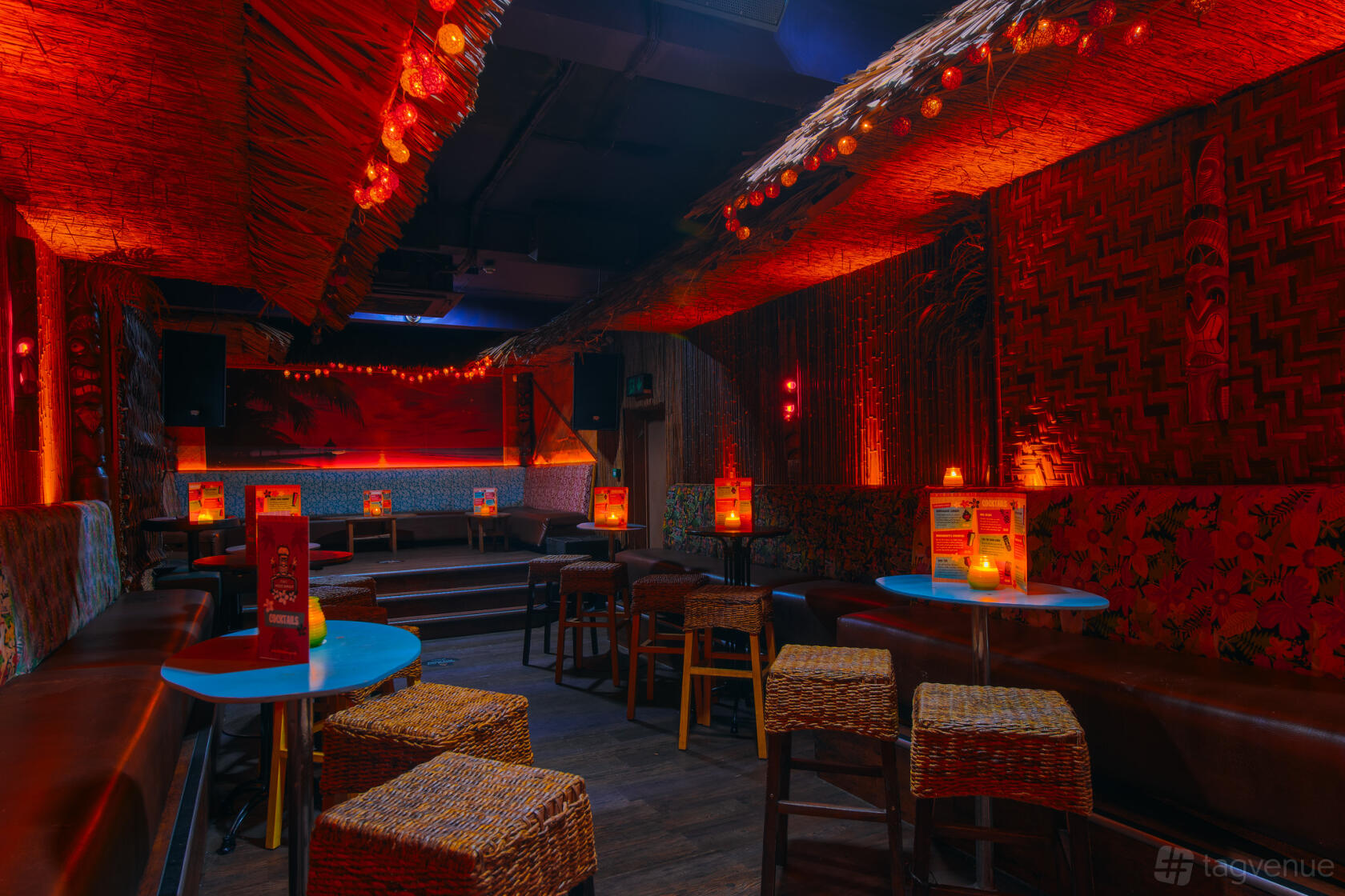 A tiki bar with woven rattan stools, blue tables, and warm orange lighting at Tiki Hideaway.