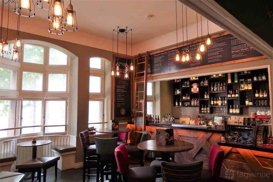 A museum lounge with large arched windows, hanging pendant lights, and a whiskey display behind the bar at Irish Whiskey Museum.