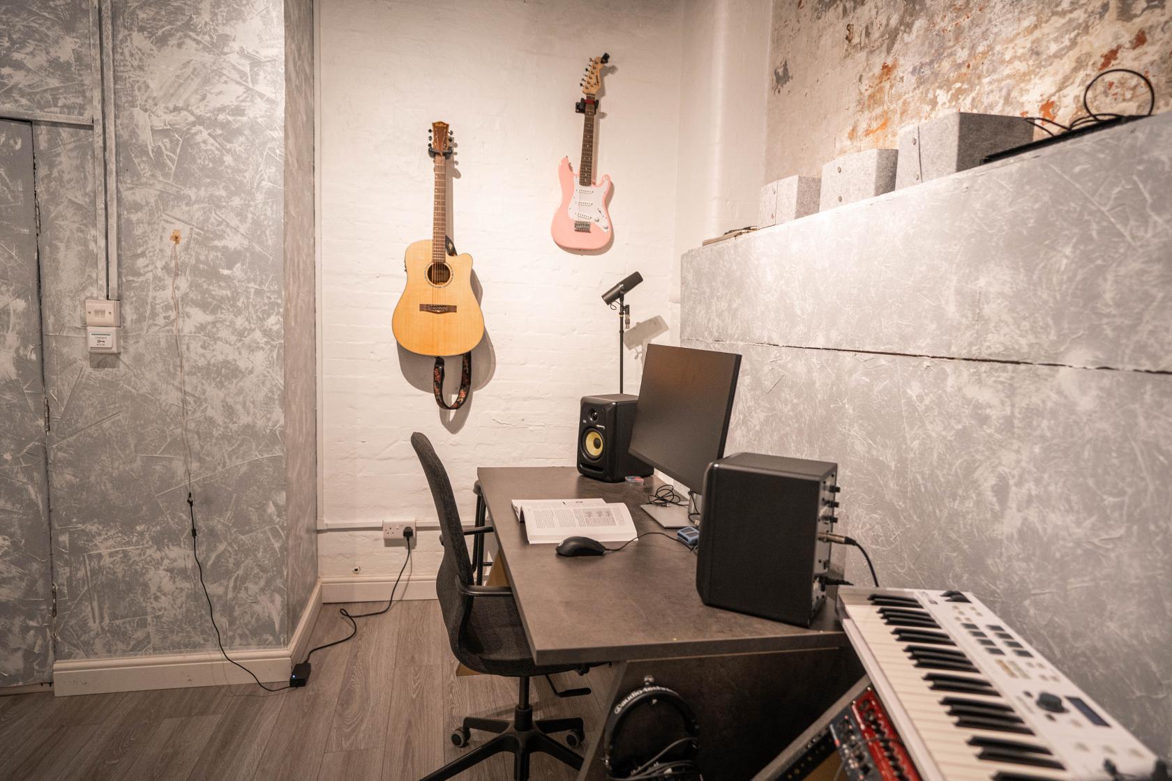 A recording studio with a workstation, studio monitors, wall-mounted guitars, and a keyboard at The Cave.