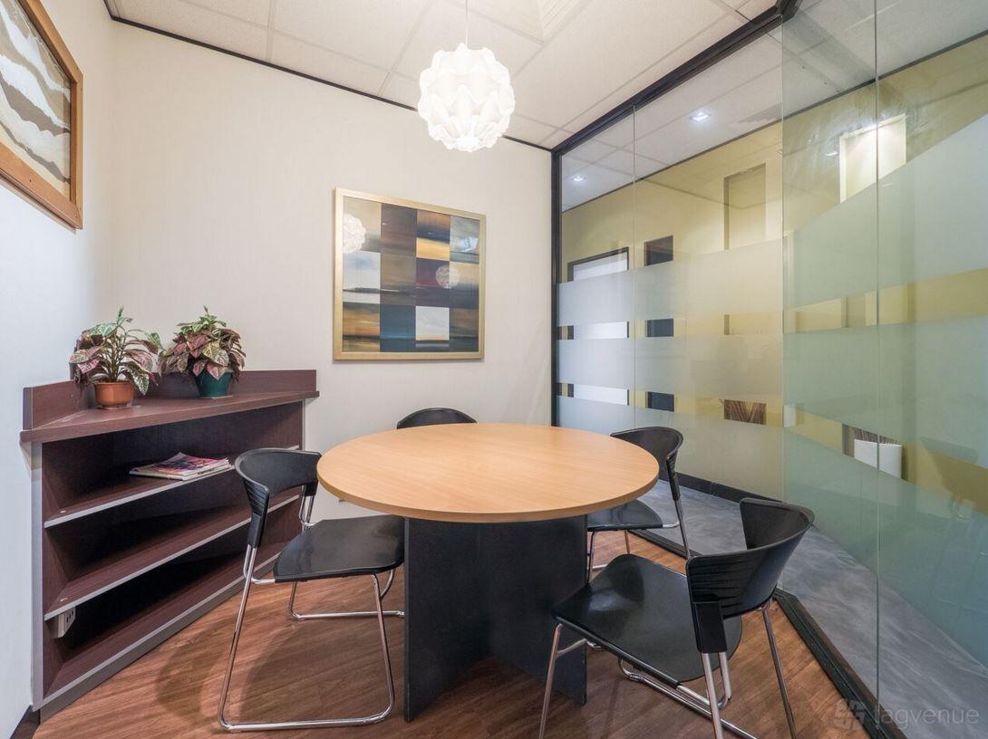 A coworking space with a round wooden table, four black chairs, frosted glass walls, and potted plants at M1 Business Centre.