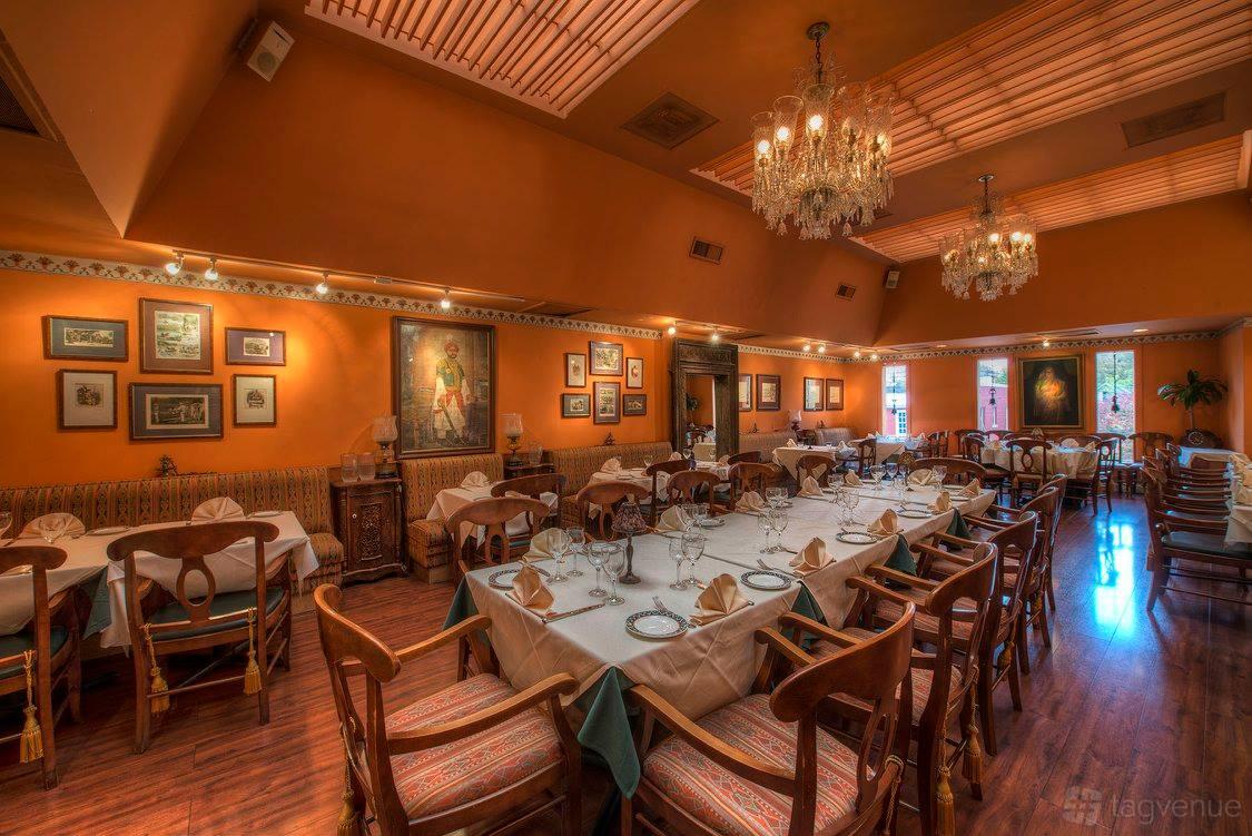 A restaurant dining room with chandeliers, framed art, and tables set with white linens at Heritage India.