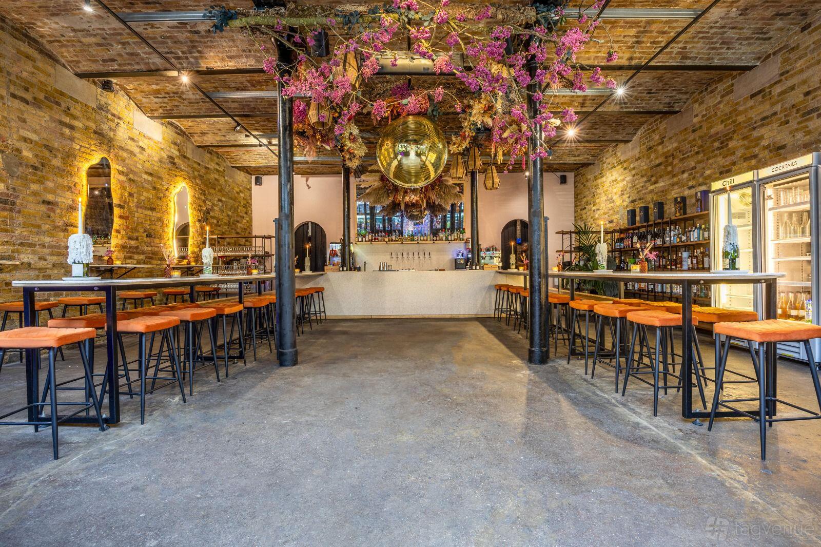 A bar with exposed brick walls, floral ceiling decor, a disco ball, and high tables with orange stools at Ester.