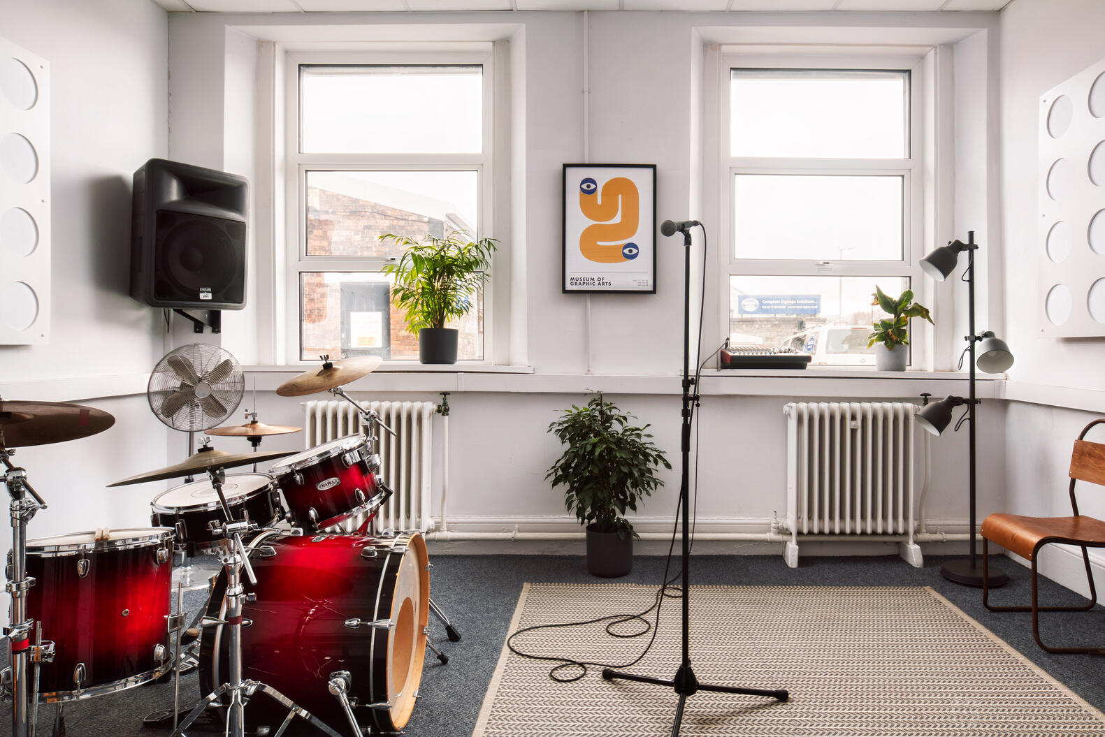 A rehearsal studio with a drum kit, microphone stand, potted plants, and large windows at Factory Studios.