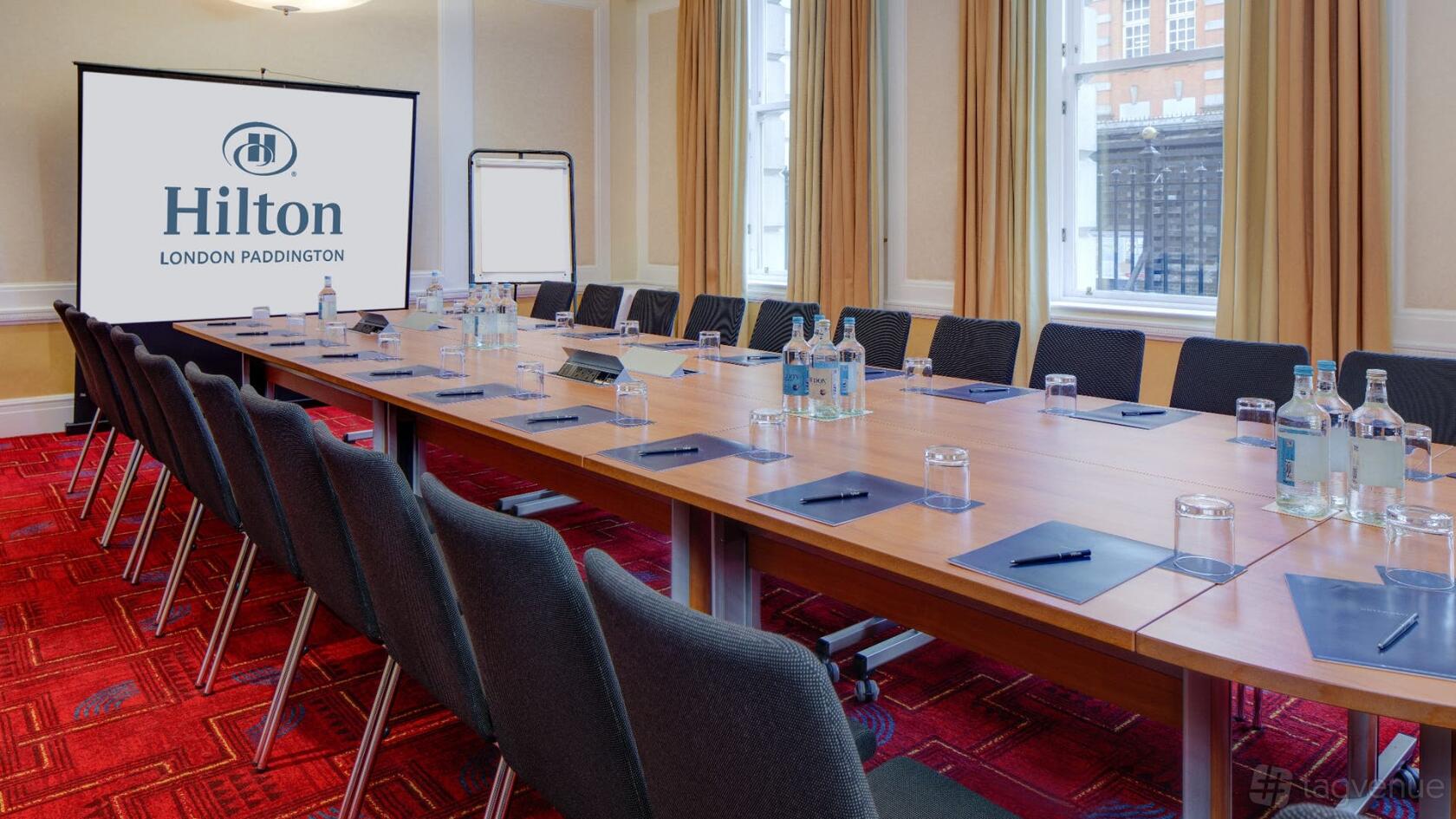 A conference room with a long boardroom table, black chairs, notepads, water bottles, and large windows at Hilton London Paddington.