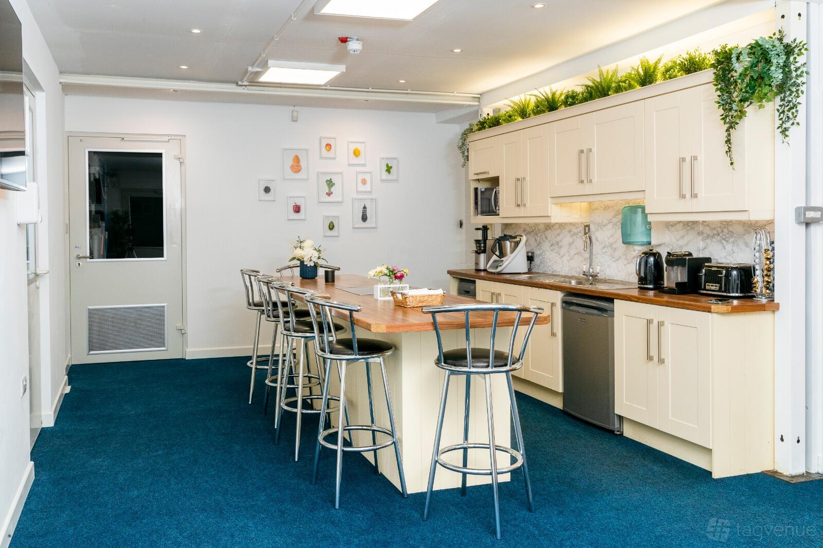A kitchen studio with a long wooden island, metal barstools, marble backsplash, and potted plants at Graco Studios.