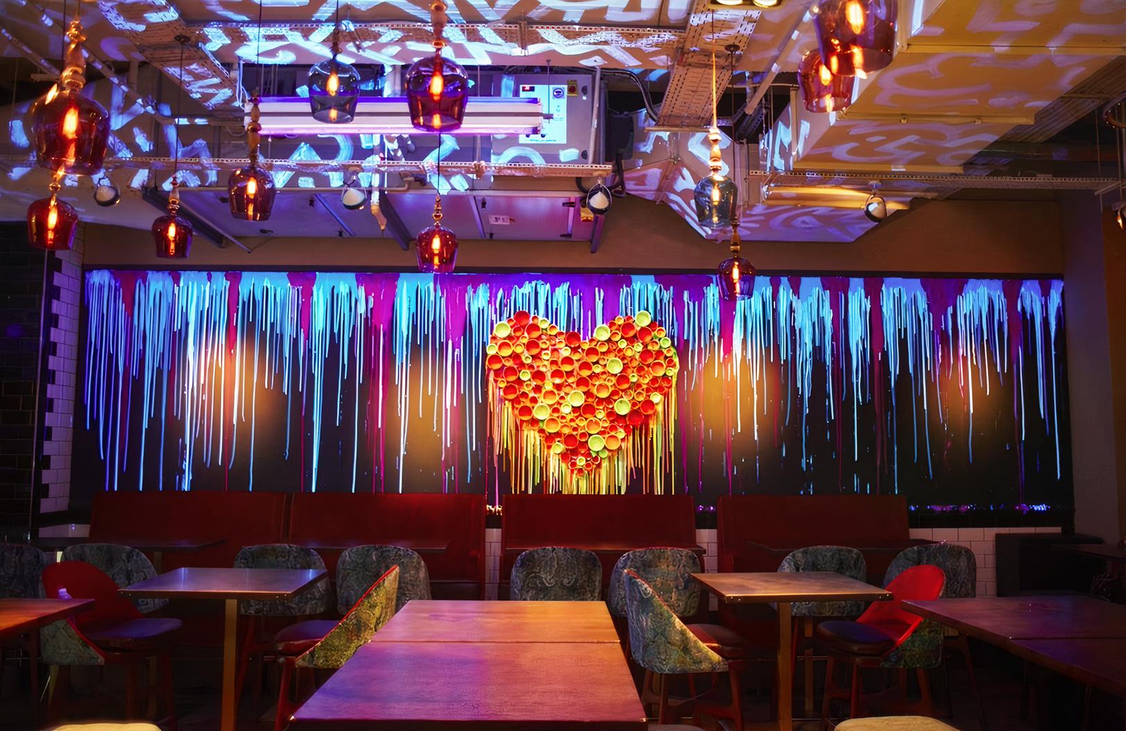A hall with eclectic lighting, velvet banquette seating, and a heart-shaped wall art installation at Bounce Farringdon.