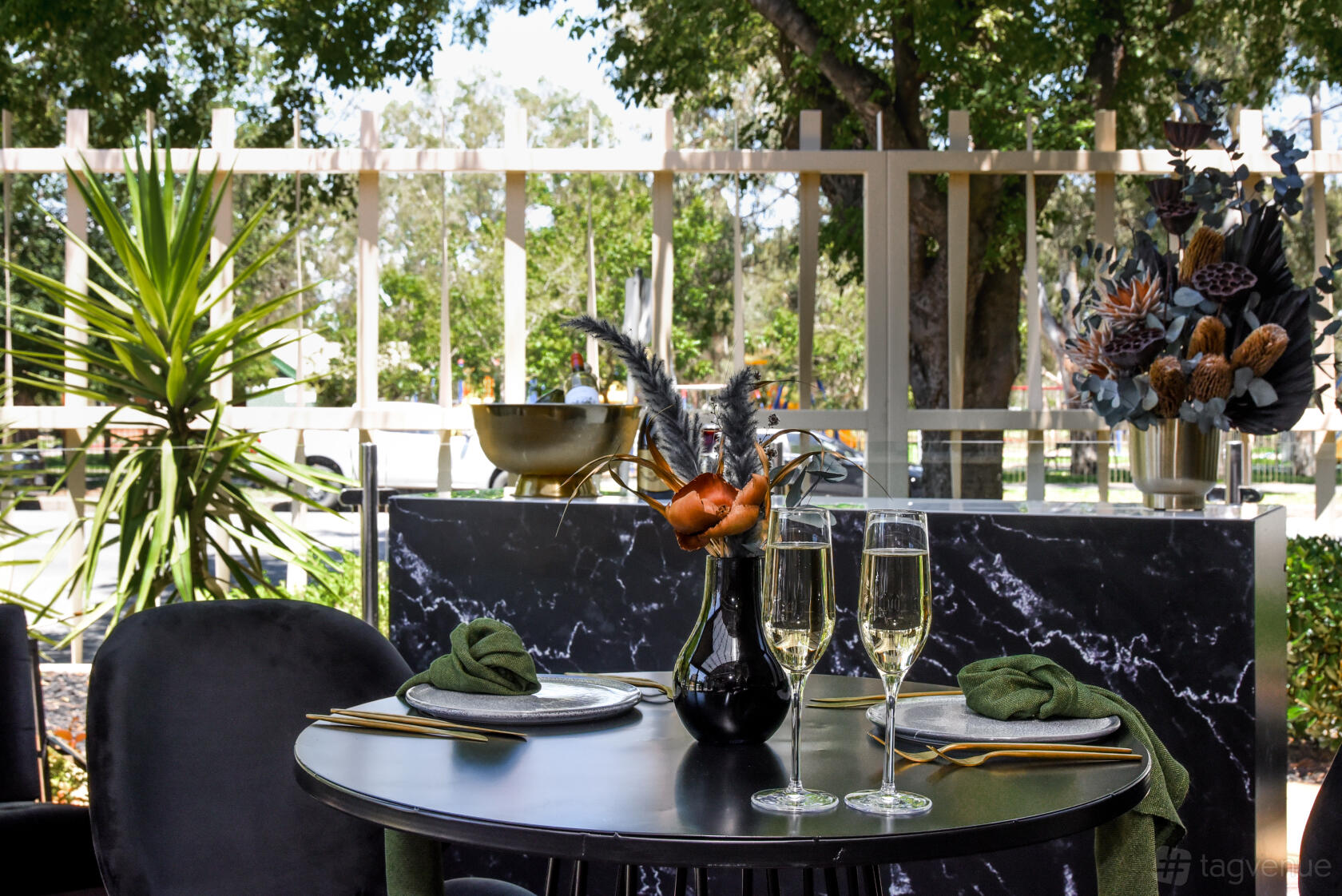 An outdoor hotel space with a small round table set for two, black marble backdrop, and leafy trees at The Terrace Hotel.