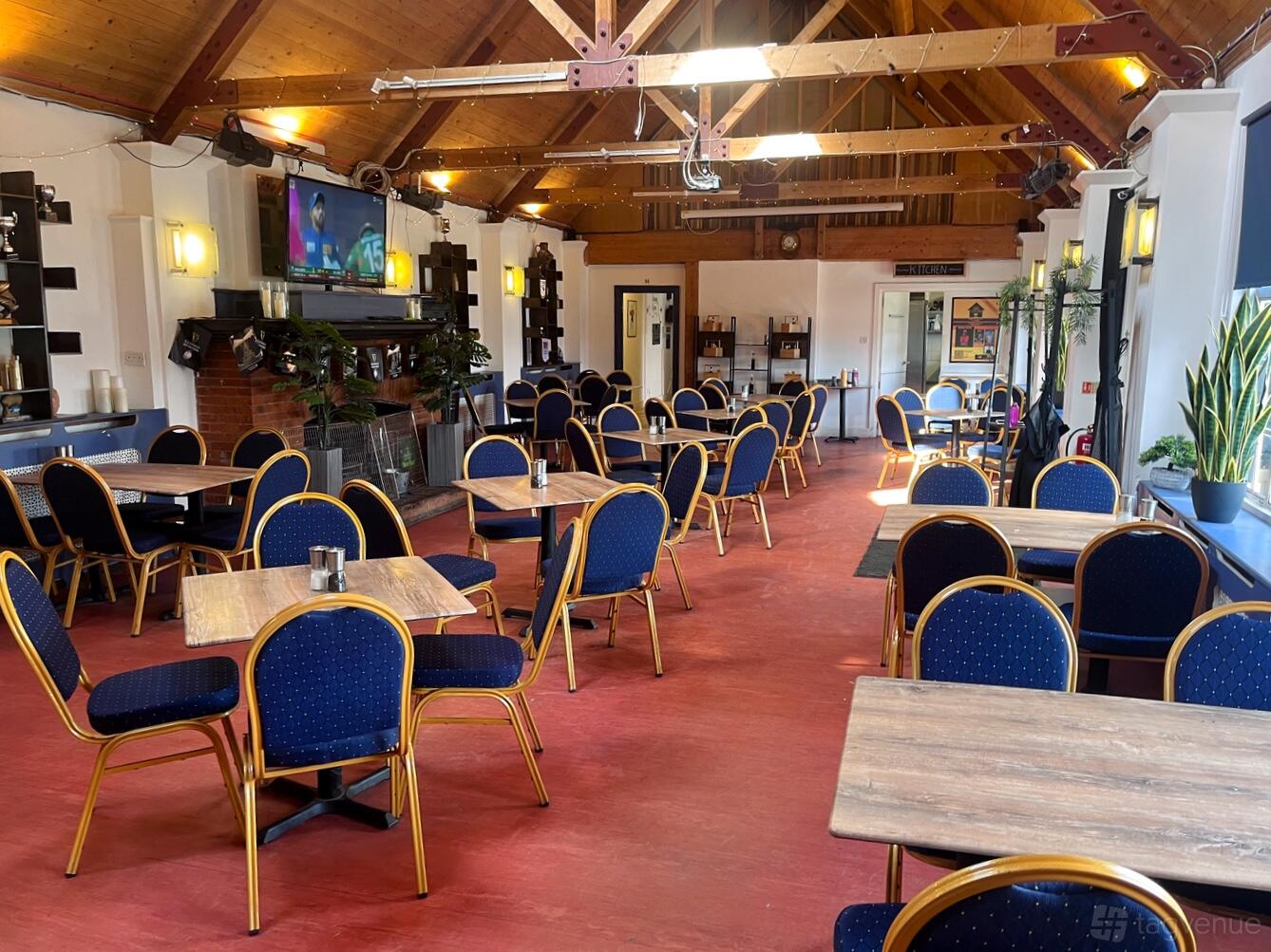 An event space with blue cushioned chairs, wooden tables, a TV, and exposed wood ceiling beams at Old Cranleighan Sports Club.
