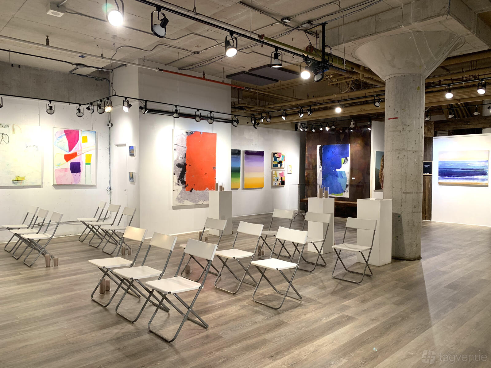 A gallery with abstract paintings, folding chairs, exposed concrete pillars and track lighting at Arta Gallery.
