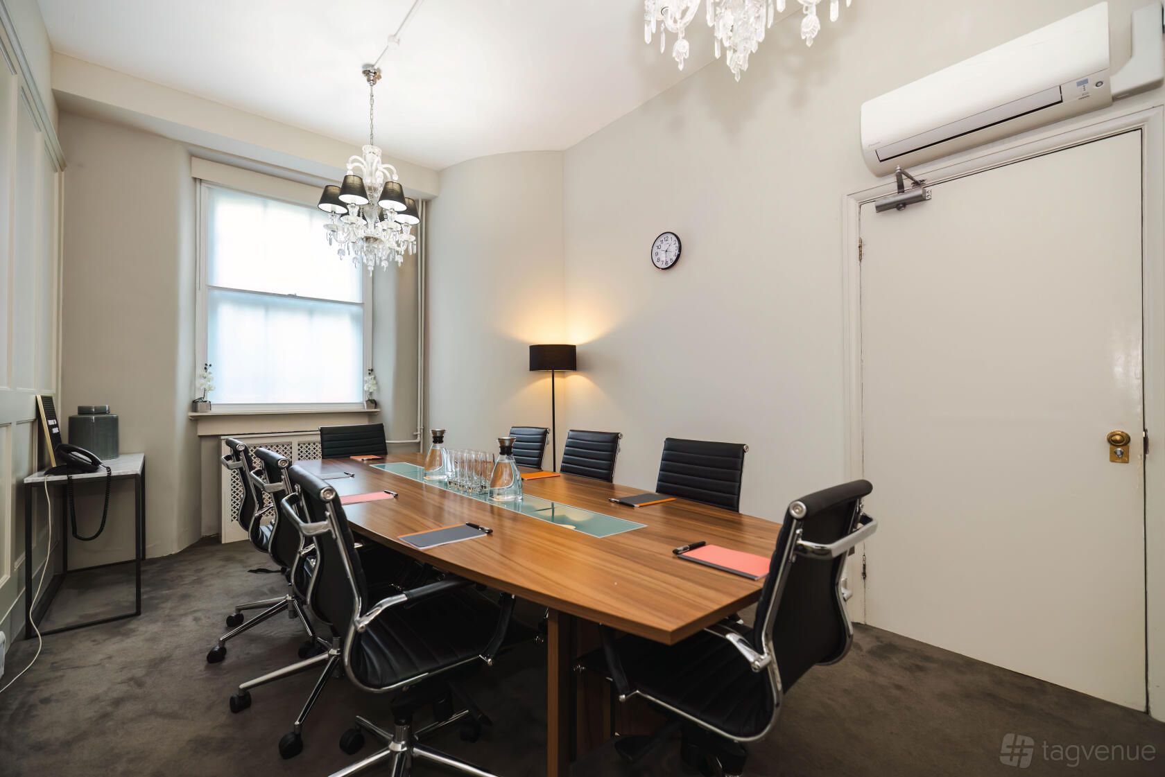 A meeting room with a wooden conference table, black leather chairs, chandeliers, and a wall clock at Grosvenor Gardens.