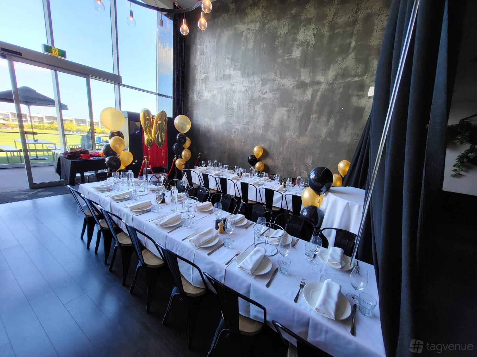 A cafe with long banquet tables covered in white linens, black chairs, and gold and black balloon decorations at The Last Piece Restaurant.