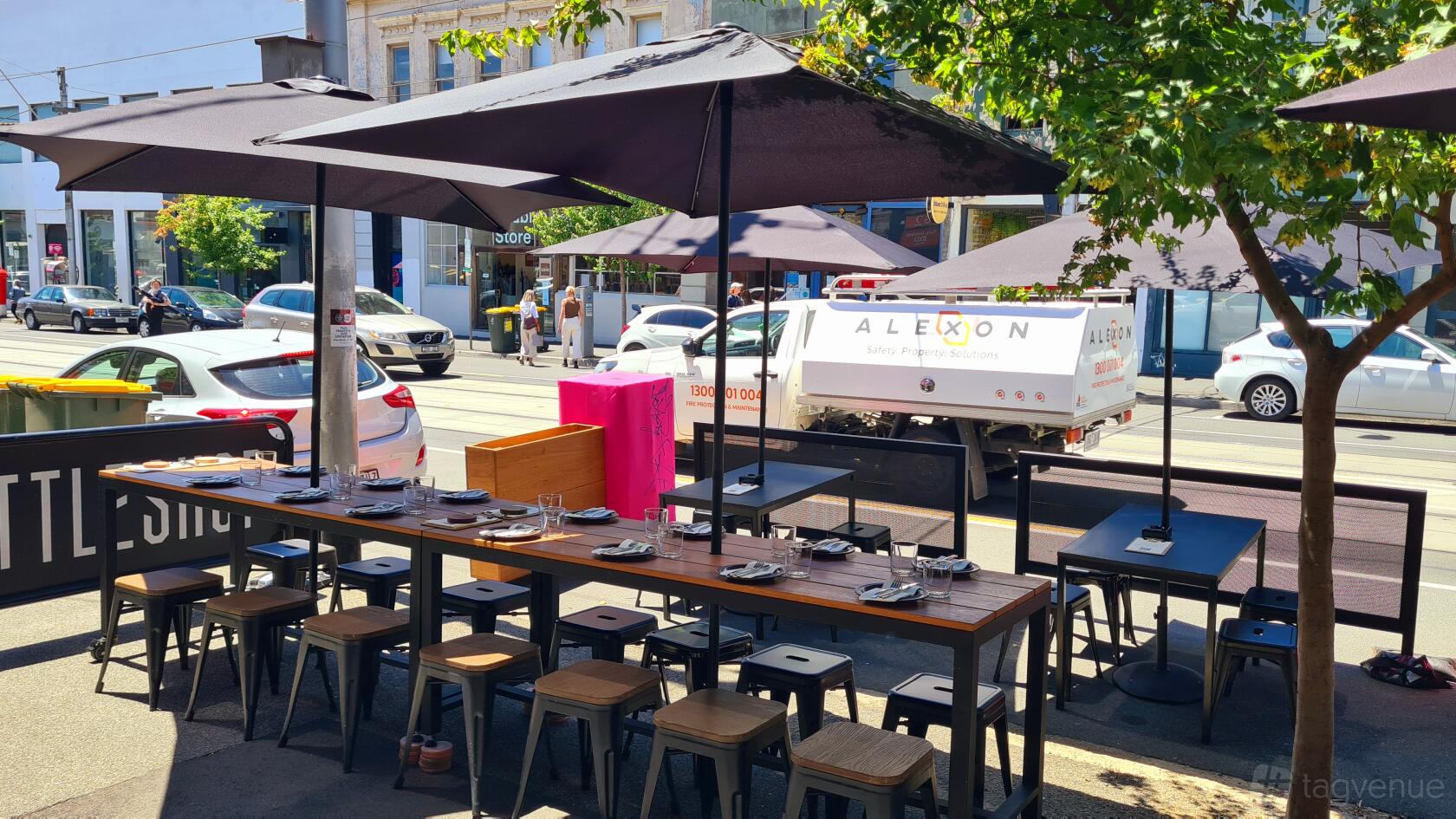 An outdoor bar area with wooden tables, metal stools, and large black umbrellas shaded by trees at Bonny.