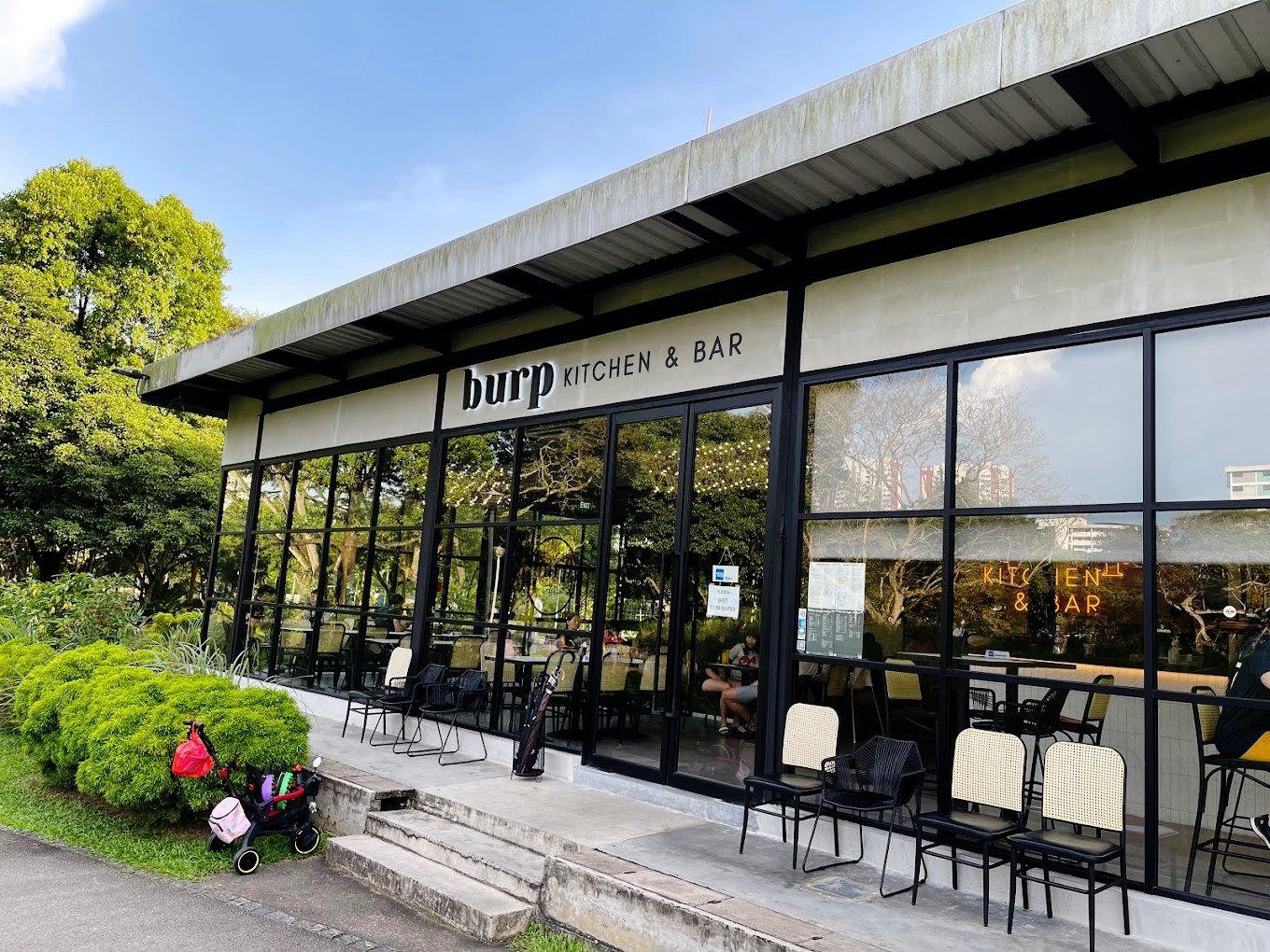 A restaurant with floor-to-ceiling glass windows, outdoor seating, and lush greenery at Burp Kitchen & Bar - Bishan Park.