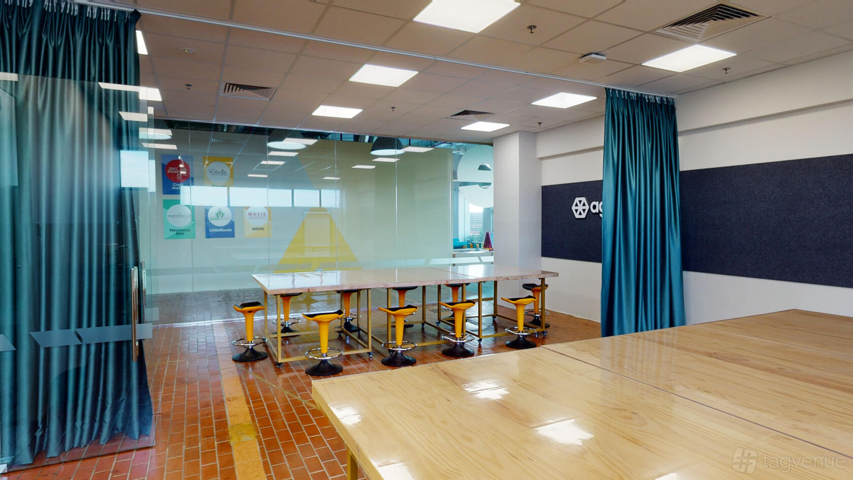 A meeting room with high tables, yellow stools, wood flooring, and glass walls at Agora Colearning.