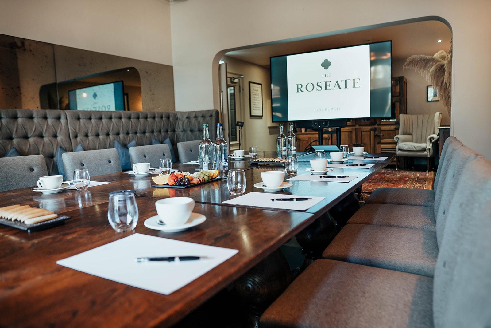A function room with a long wooden table, upholstered chairs, and a large presentation screen at The Roseate Edinburgh.