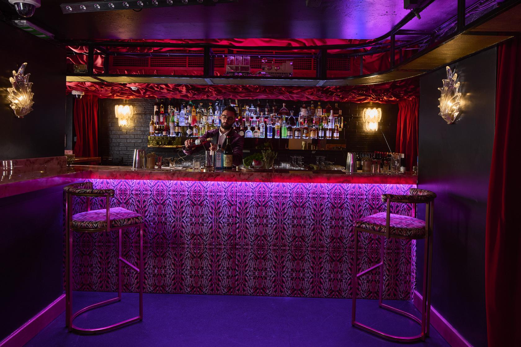 A nightclub bar with a neon-lit counter, backlit liquor shelves, and velvet barstools at The Knox.