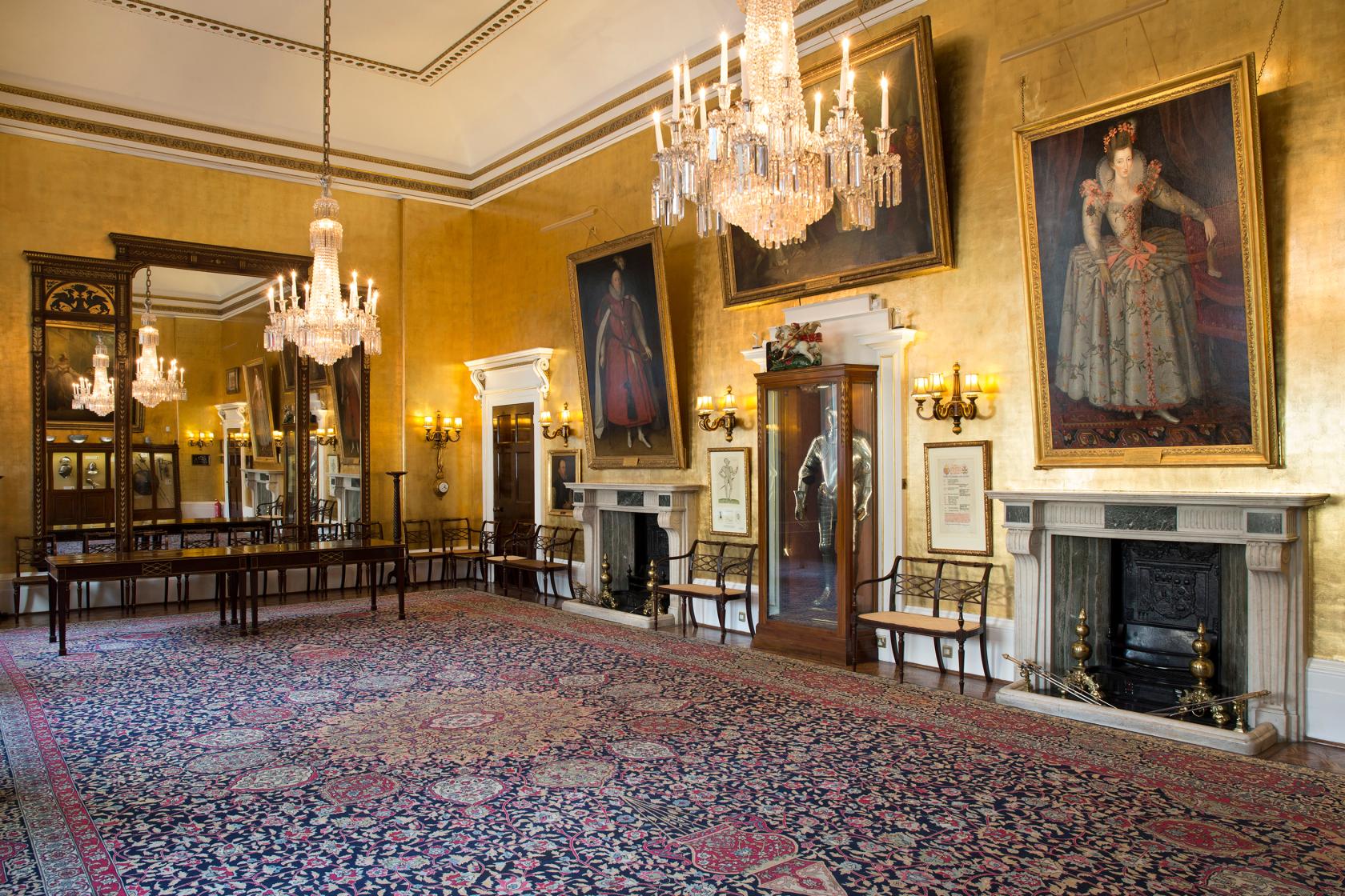 A historic event space with gold walls, chandeliers, ornate fireplaces, and large portraits at Armourers' Hall Gold Drawing Room.