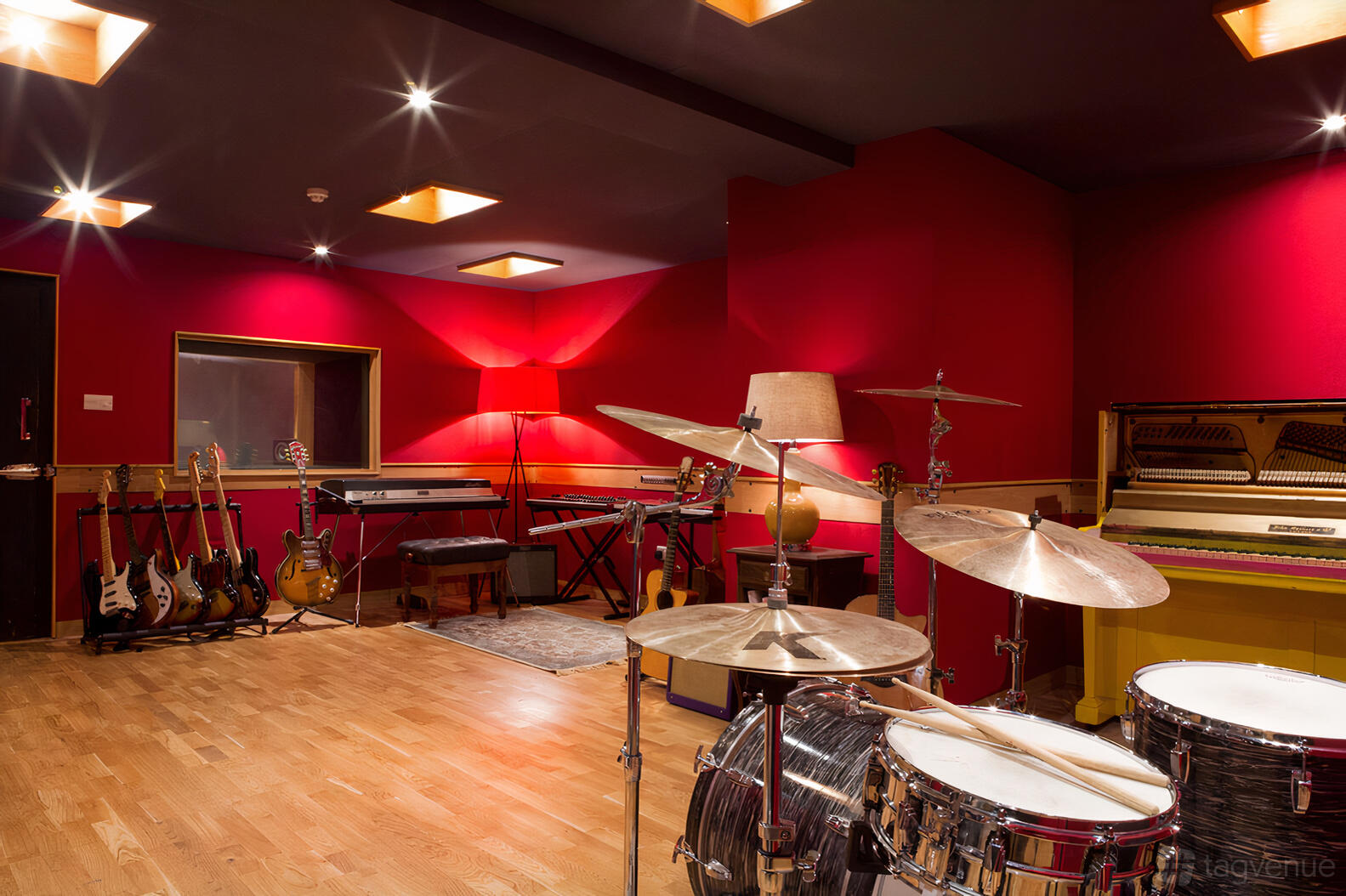 A recording studio with red walls, a drum set, upright piano, electric guitars, and hardwood floors at Dock Street Studios.