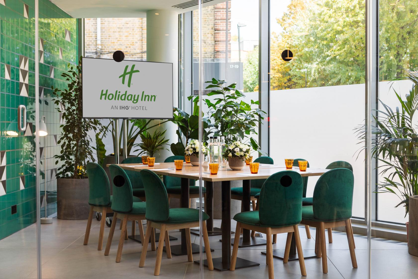A meeting room with green velvet chairs, wood table, potted plants, and floor-to-ceiling windows at Holiday Inn London Whitechapel.