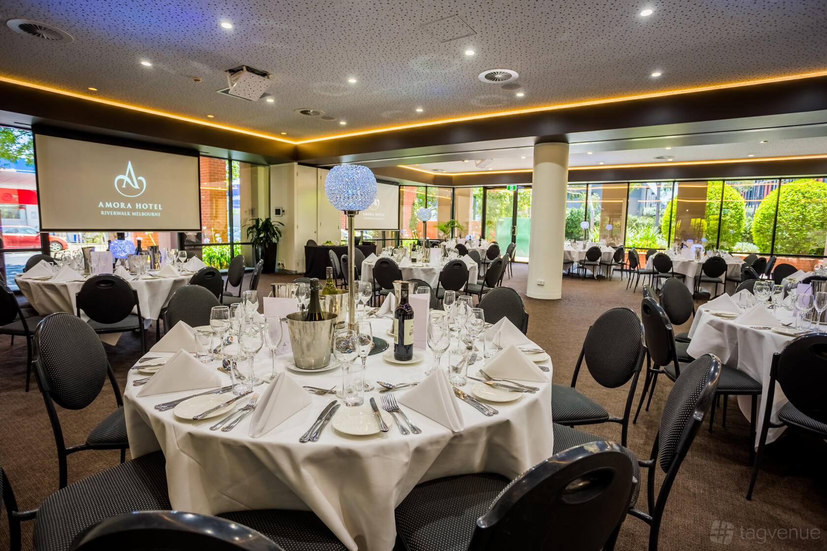 An event space in a hotel with round tables set with white linens, glassware, and floor-to-ceiling windows at Amora Hotel Riverwalk Melbourne.
