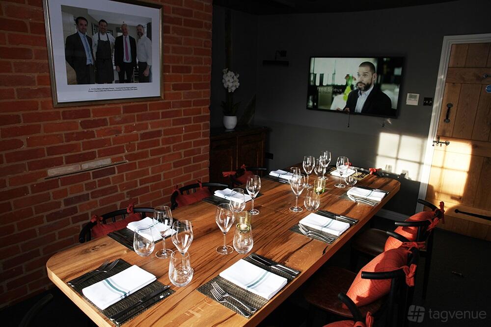 A dining room with a long wooden table, set with glassware and napkins, exposed brick wall, and wall-mounted TV at Galvin Green Man.