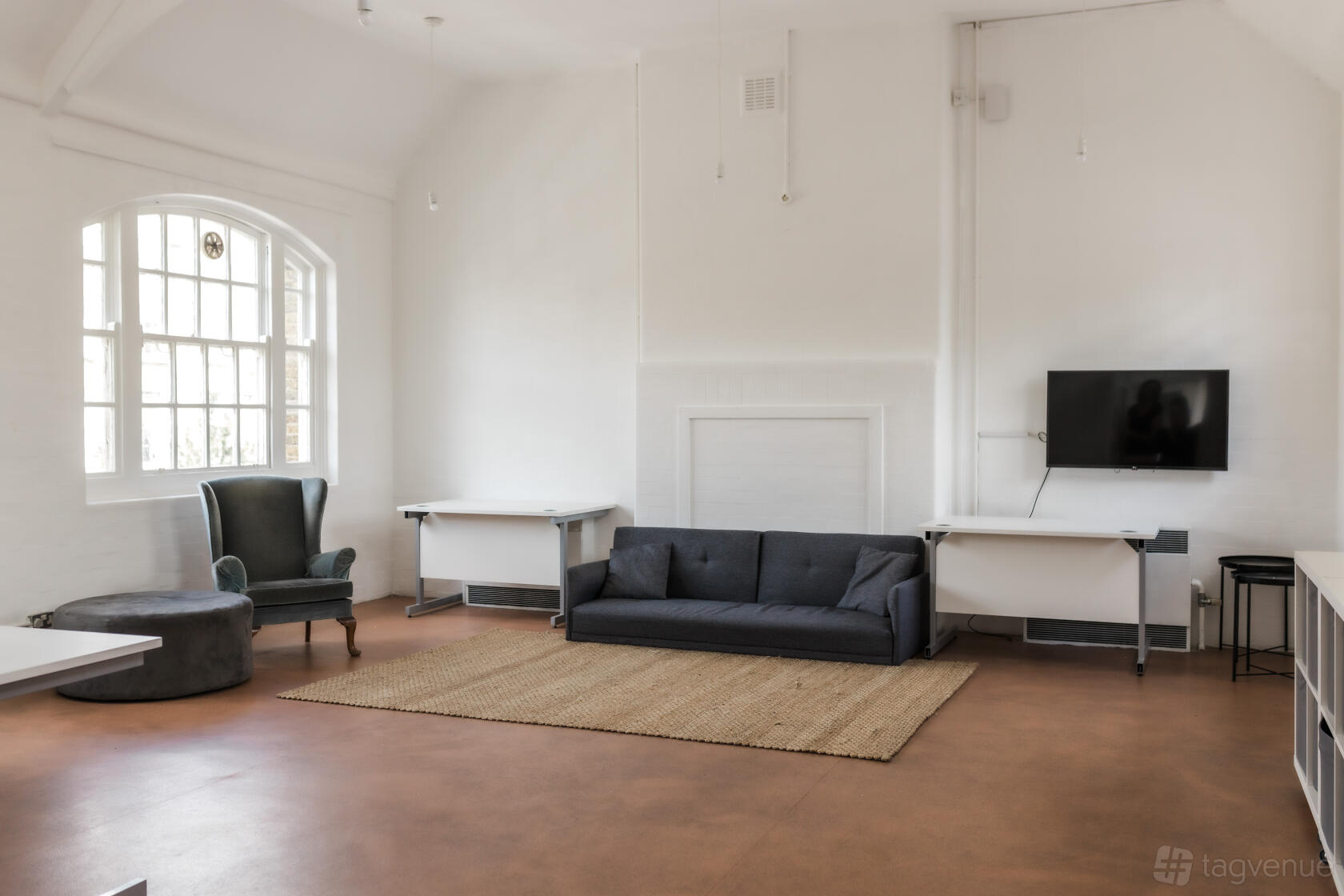 A lounge with a dark sofa, armchair, wall-mounted TV, large arched window, and neutral rug at 1-3 Coborn Street.