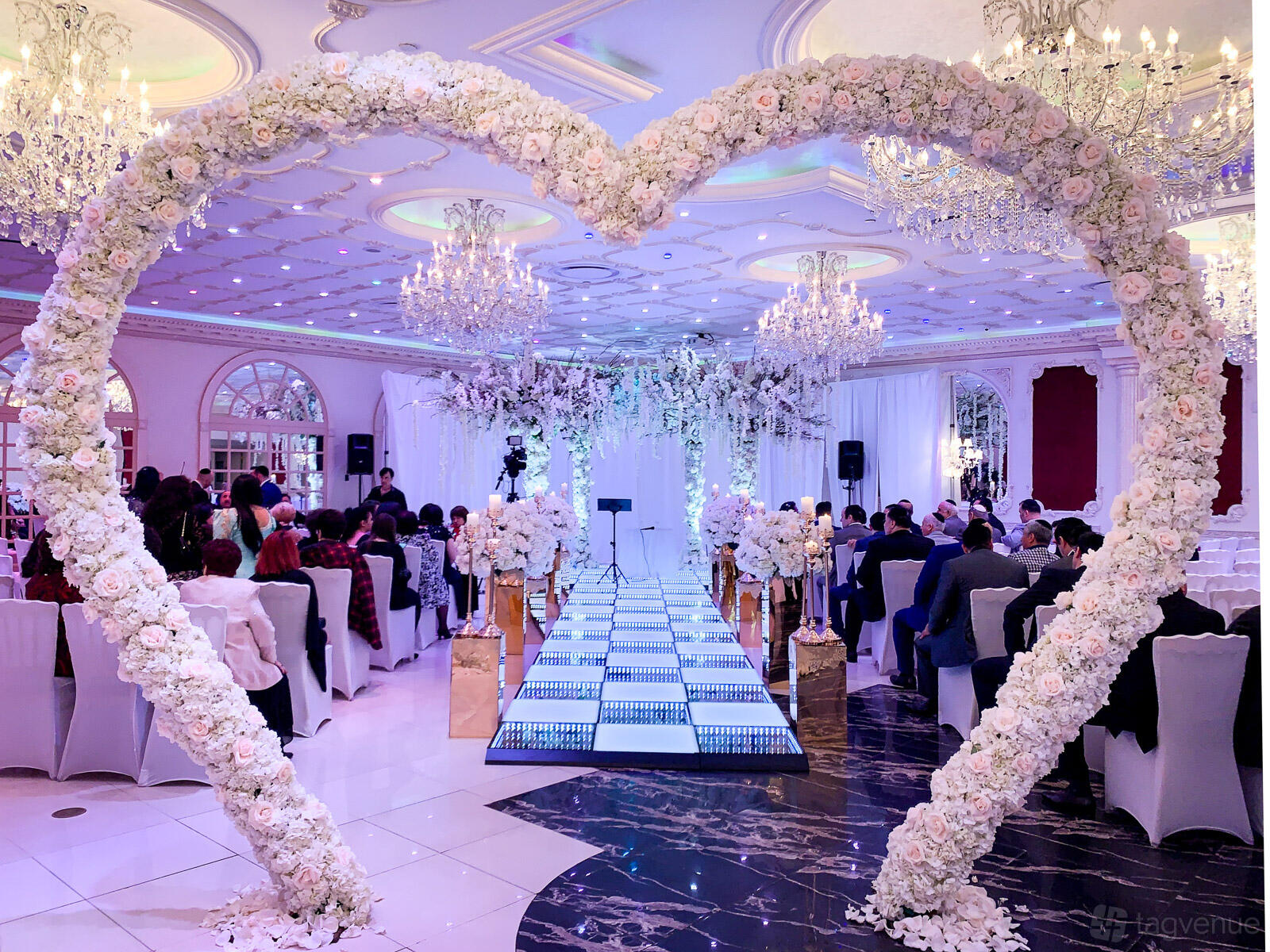 An event venue with a floral heart arch, aisle lined with white flowers, and crystal chandeliers at Da Mikele Illagio.