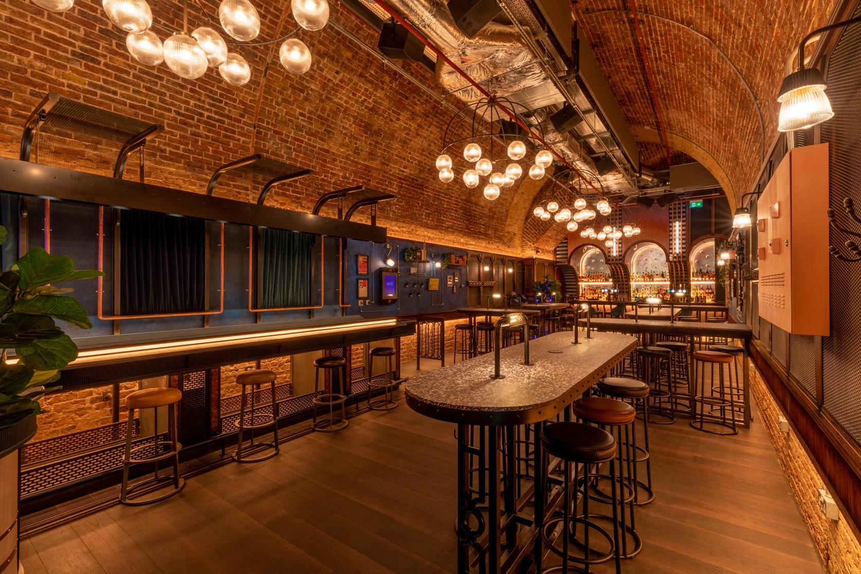 A cocktail bar with exposed brick arches, globe pendant lights, and high tables at Electric Shuffle London Bridge.