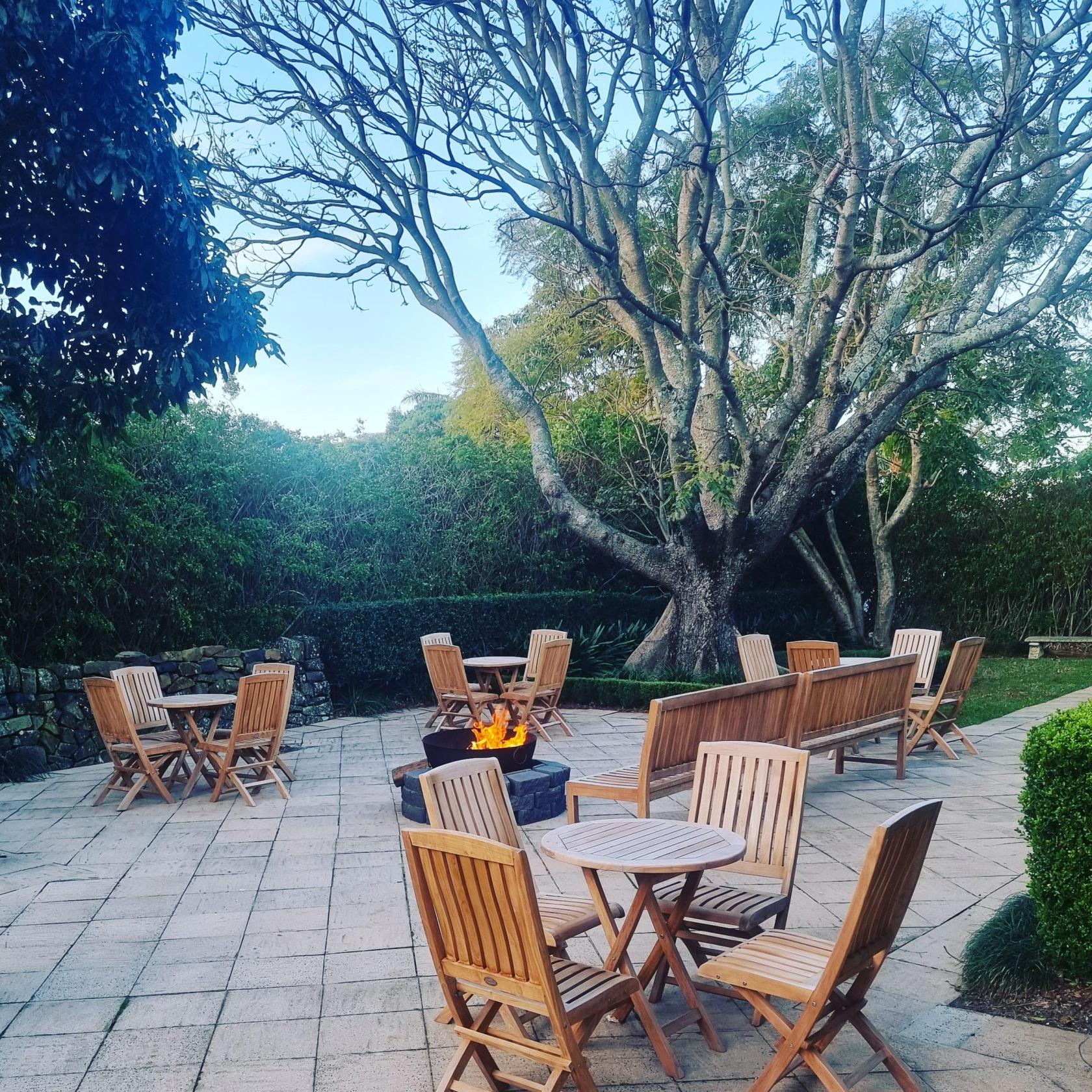 An outdoor hotel patio with wooden tables, chairs, and a fire pit under tall trees at Mercure Gerringong Resort.