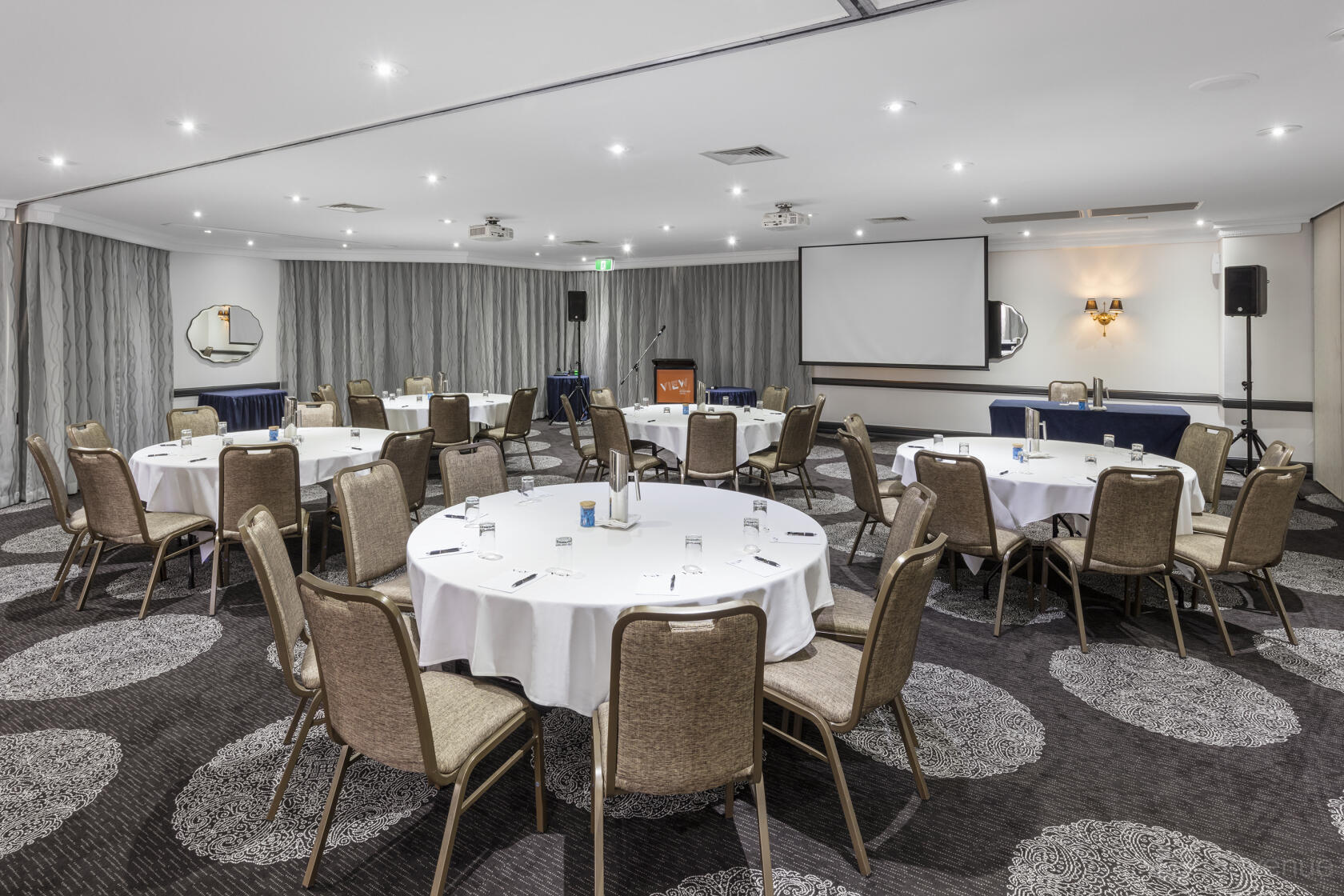 A function room with round tables, beige chairs, patterned carpet, and a projector screen at View Brisbane.