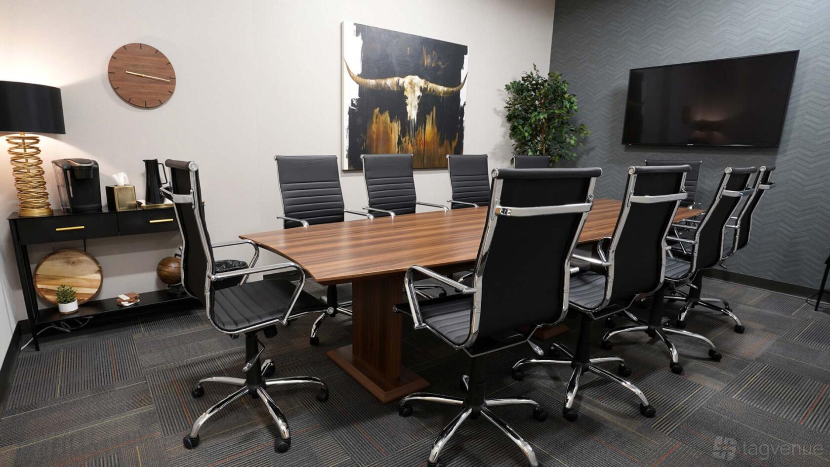 A boardroom with a long wooden table, black leather chairs, wall art, and a large display screen at Executive Workspace Frisco Station.