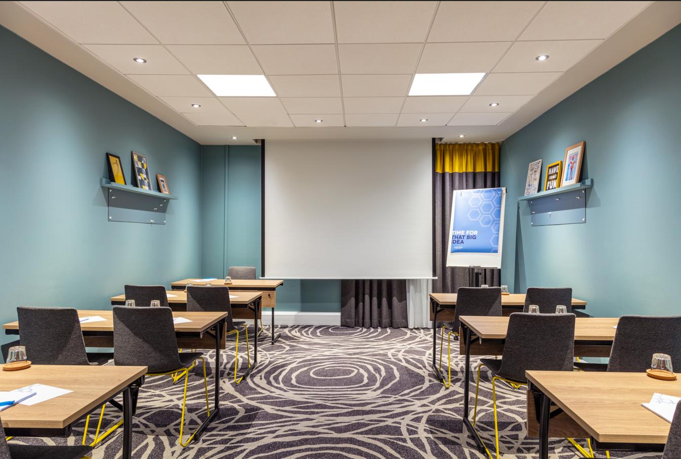 A meeting room with patterned carpet, framed art on shelves, and a projector screen at Novotel Edinburgh Centre.